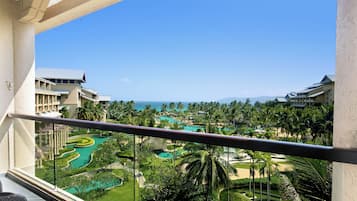 A resort balcony with a view of a landscaped pool area, palm trees, and a distant ocean.