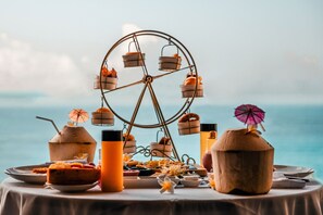 Une table dressée avec une variété de plats et de boissons, une roue décorative et une vue sur l’océan.