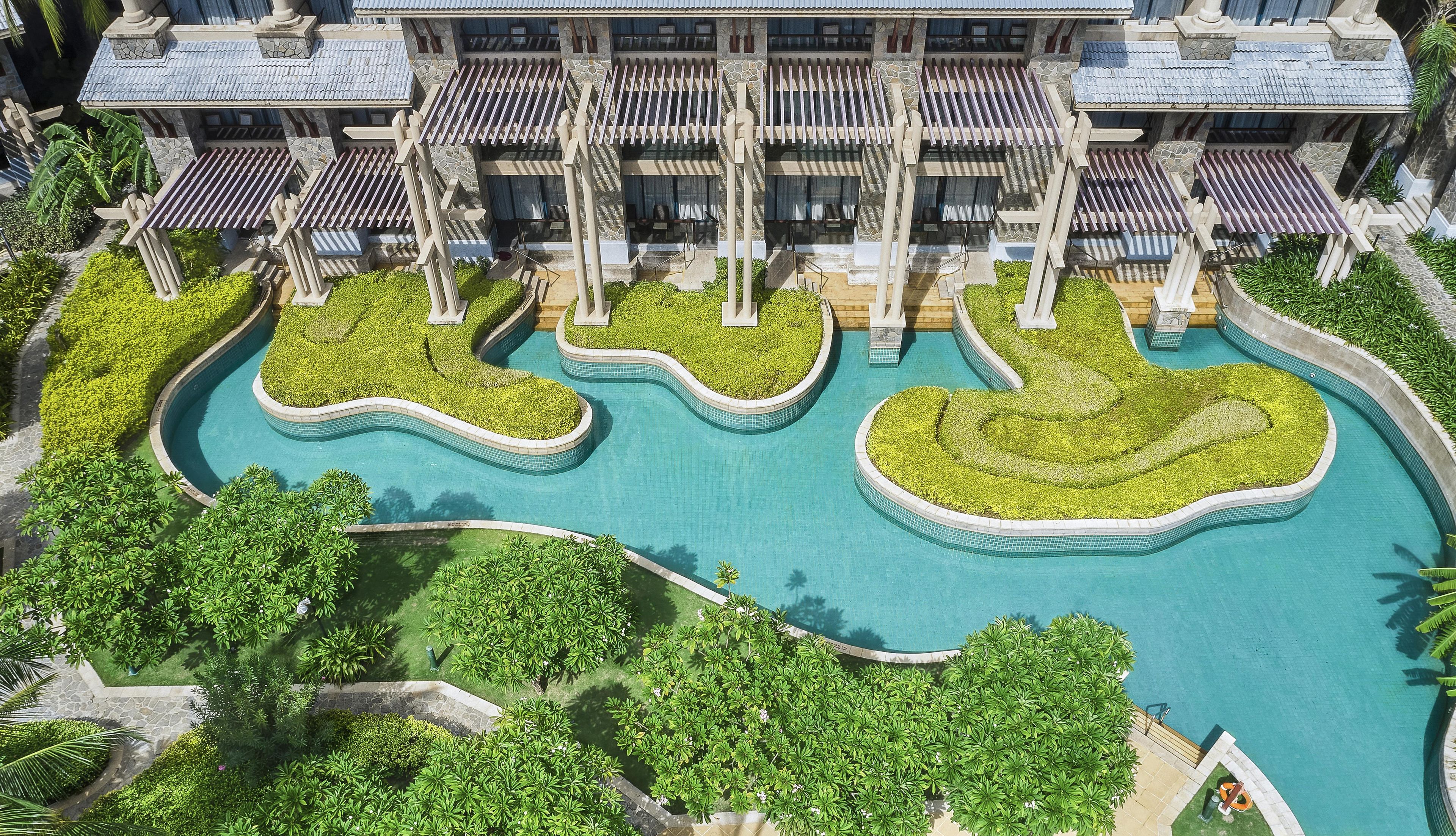 A pool with multiple sections, surrounded by lush greenery and a building with balconies.