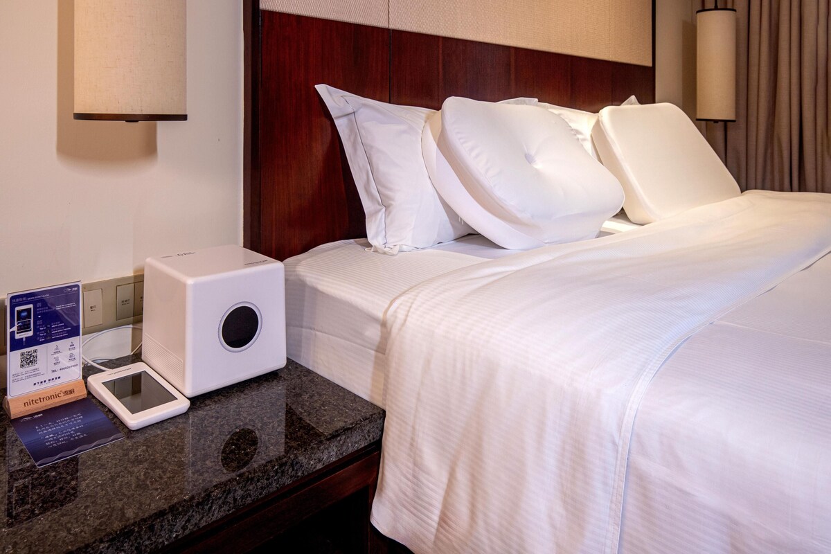 Une chambre d’hôtel avec un lit parfaitement fait, une table de chevet sur laquelle se trouvent un haut-parleur blanc et une tablette, et une tête de lit en bois.