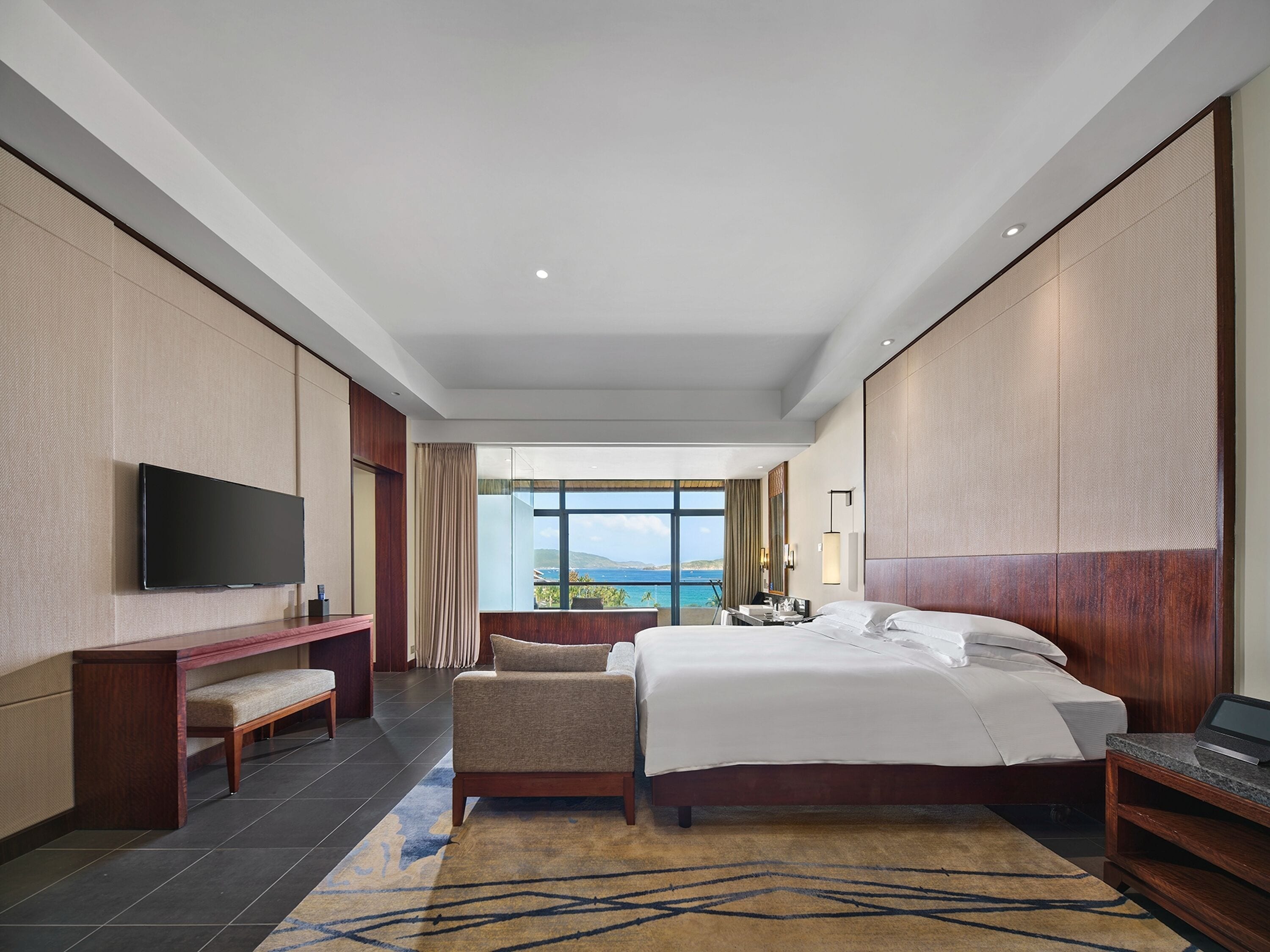 A hotel room with a large bed, a TV, a desk, and a view of the sea.