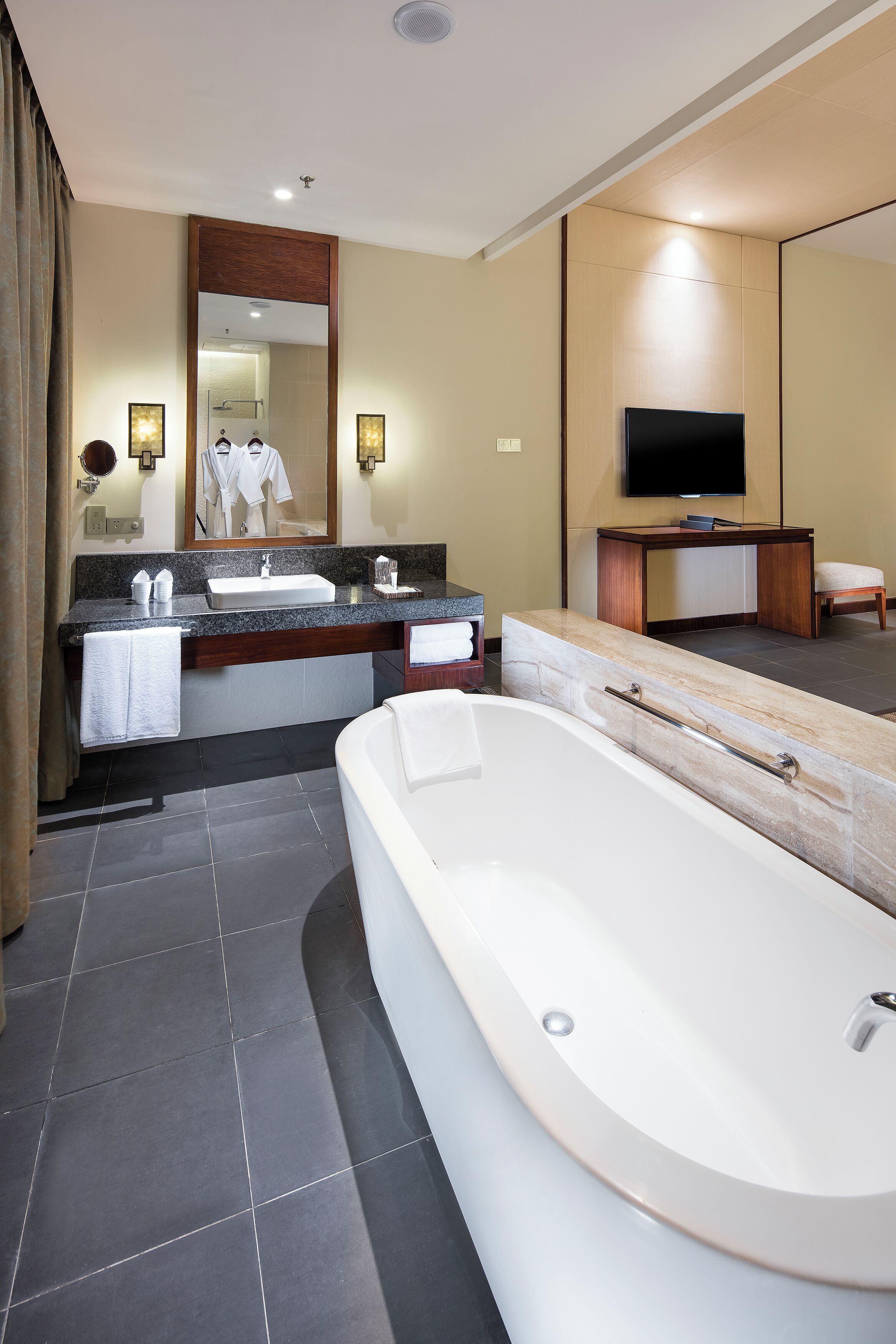 A modern bathroom with a large bathtub, a vanity with a mirror, and a television mounted on the wall.