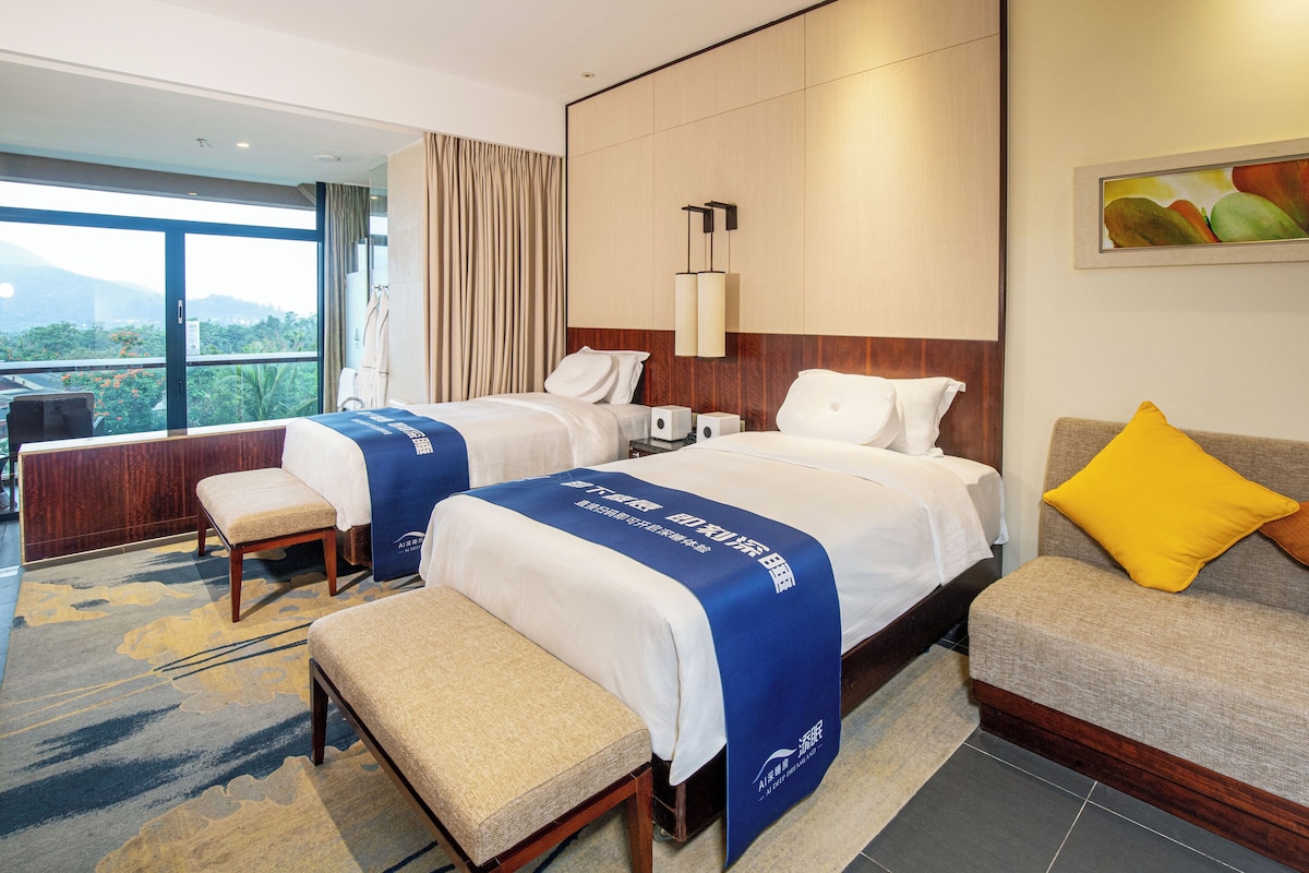 A hotel room with two beds, a sofa, a bench, and a large window offering a view of greenery.