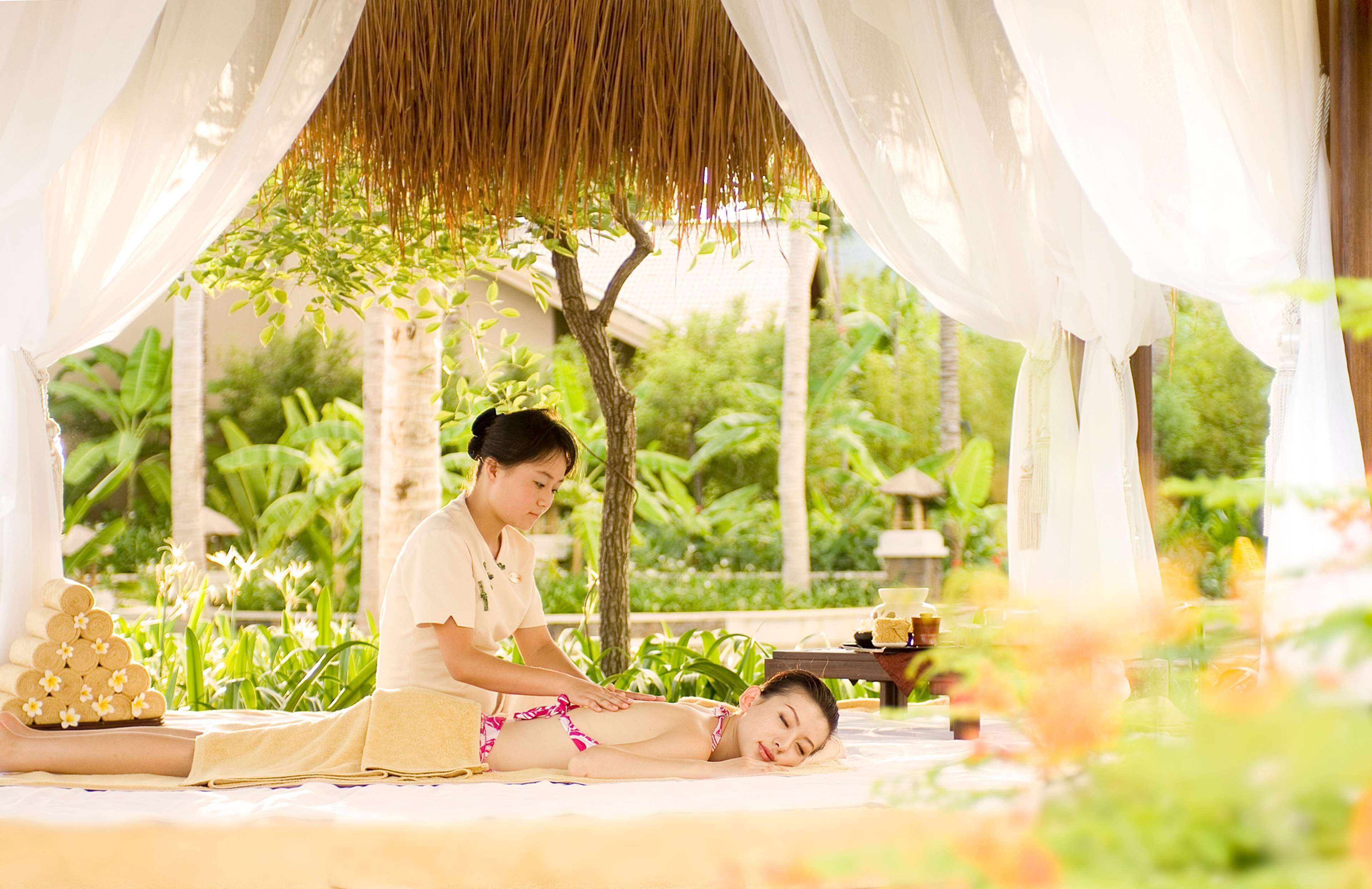 A person receiving a massage in a serene outdoor setting with lush greenery and a thatched roof structure.