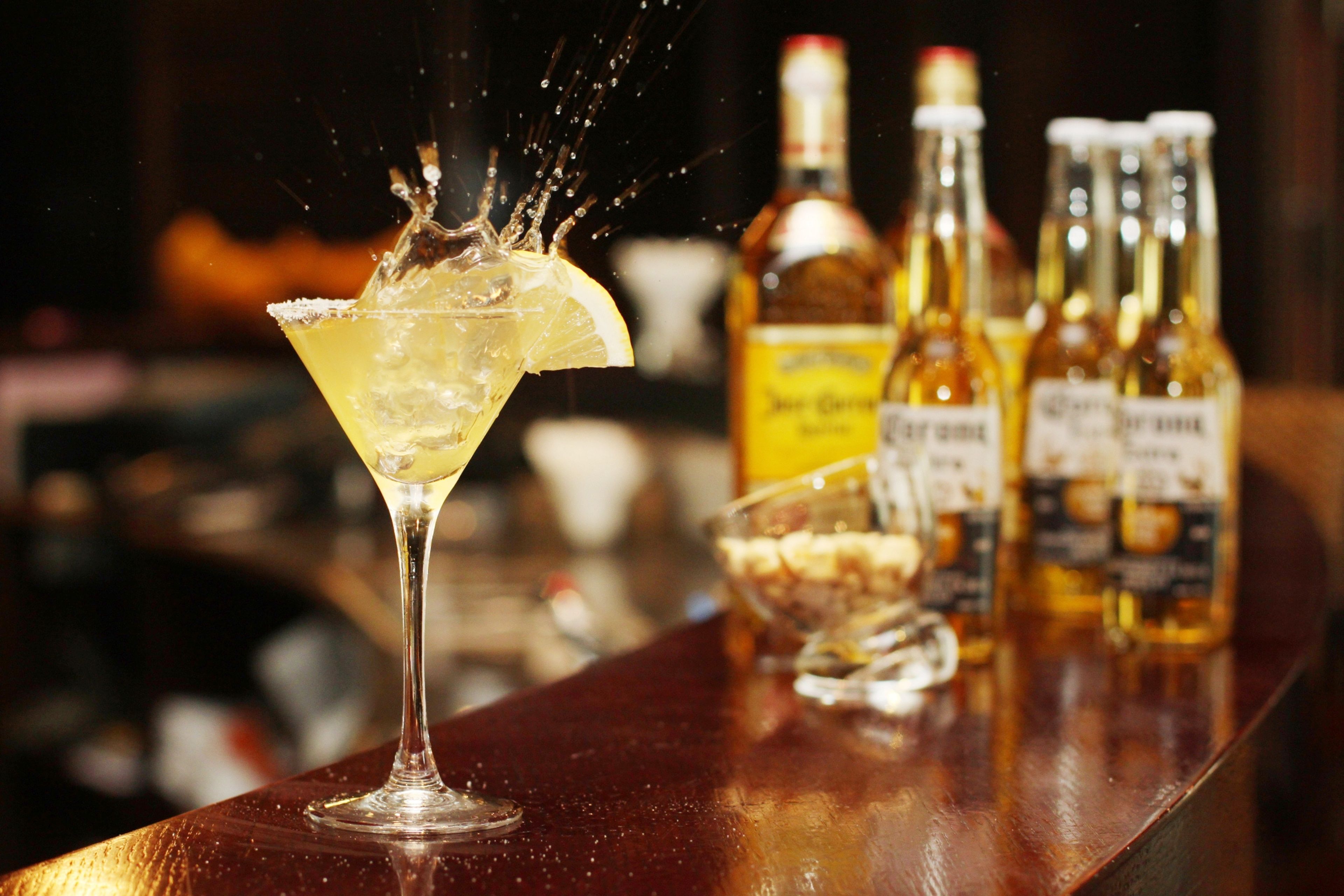 A cocktail with a lemon slice on the rim, a bottle of whiskey, and several bottles of liquor on a bar counter.