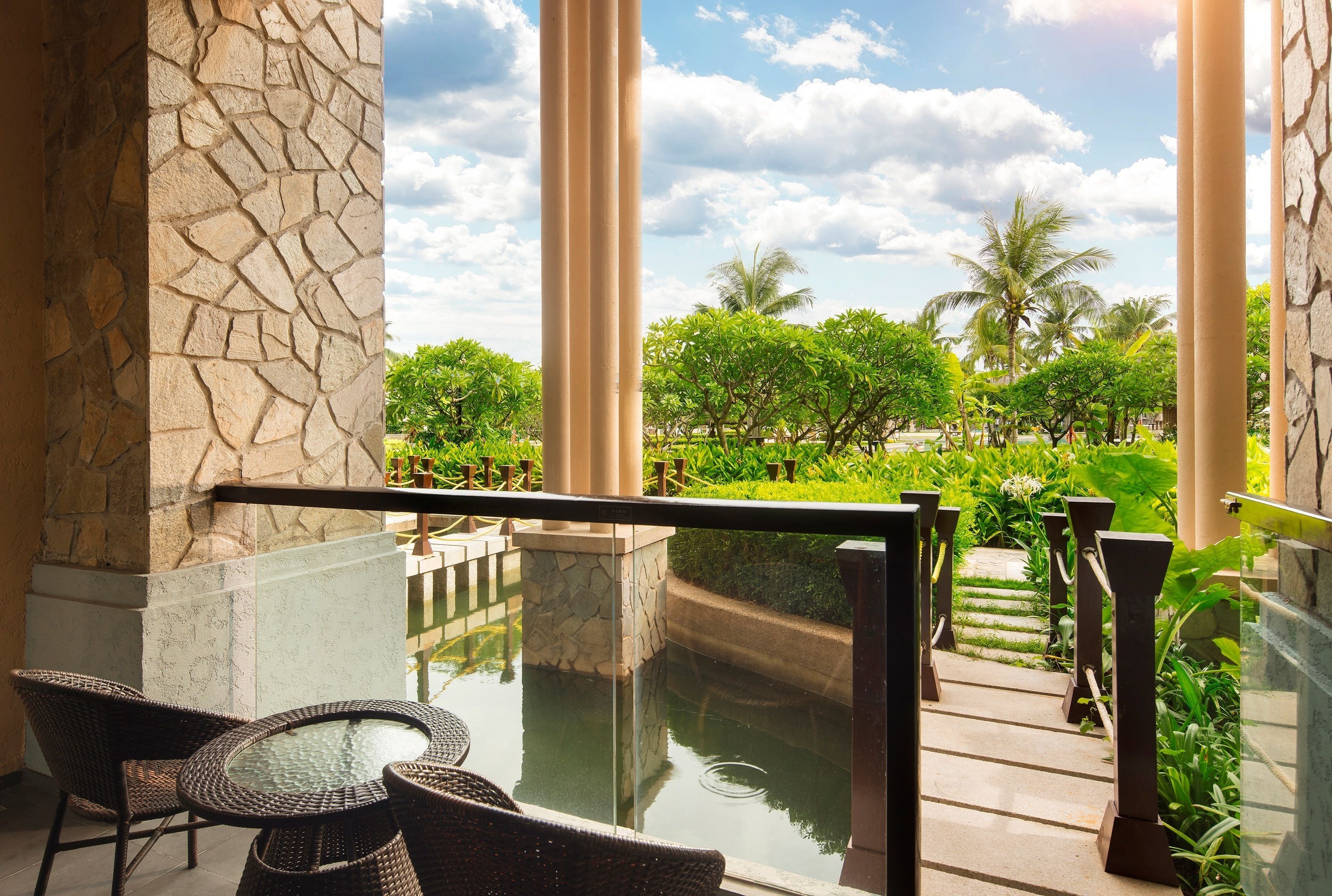 A glass-enclosed outdoor area with a stone wall, a small pool, wicker furniture, and a view of a garden with palm trees.