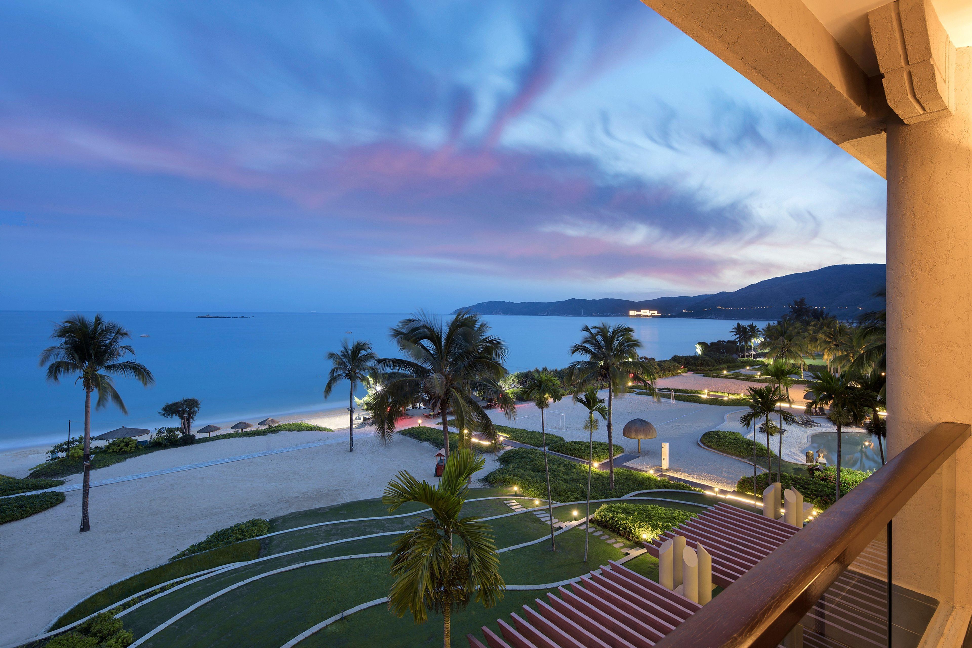 A beachfront property with a view of the sea, palm trees, and a well-lit pathway.
