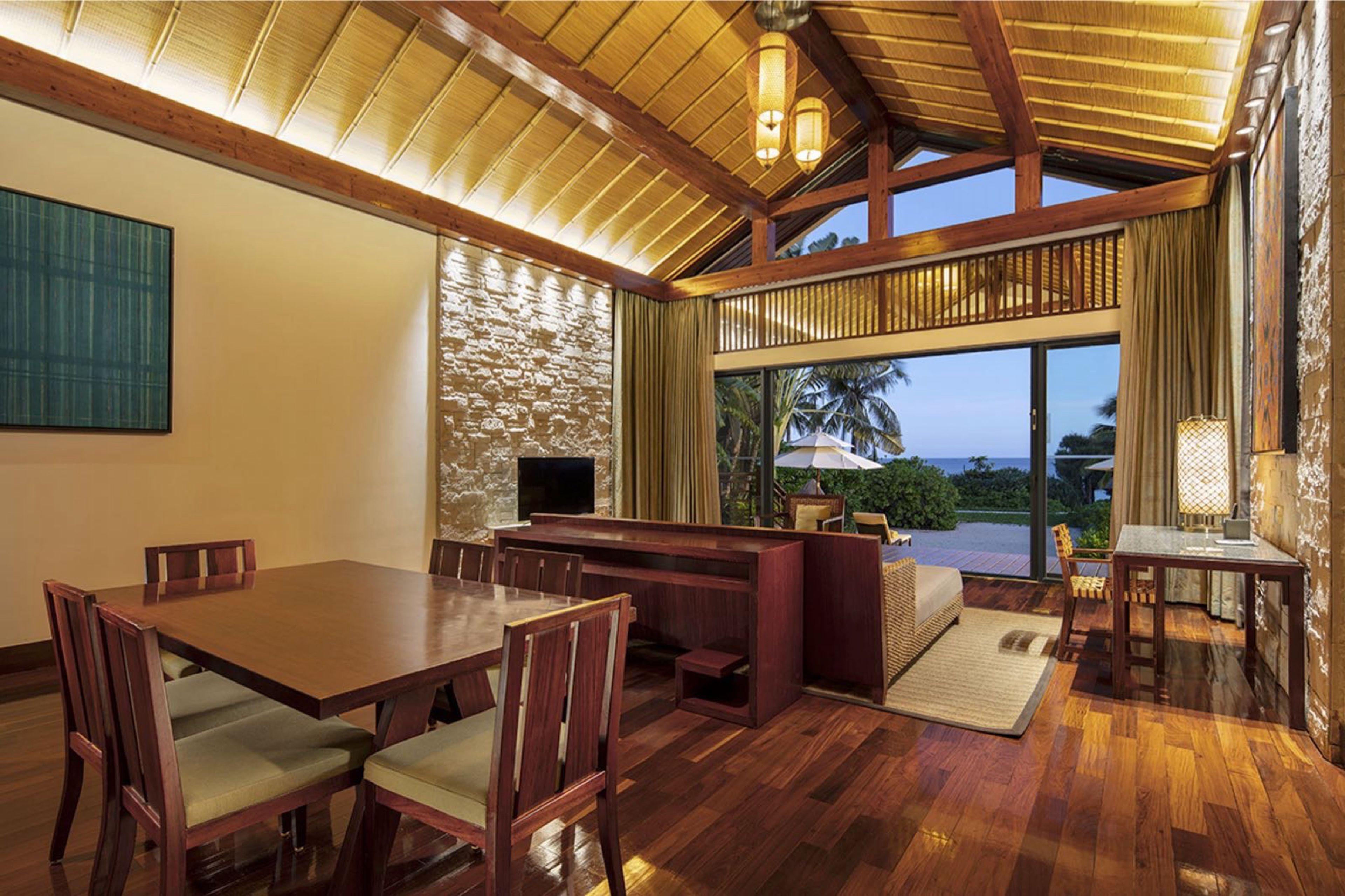Uma ampla sala de jantar com mesa e cadeiras de madeira, parede decorativa em pedra e vista para o exterior.