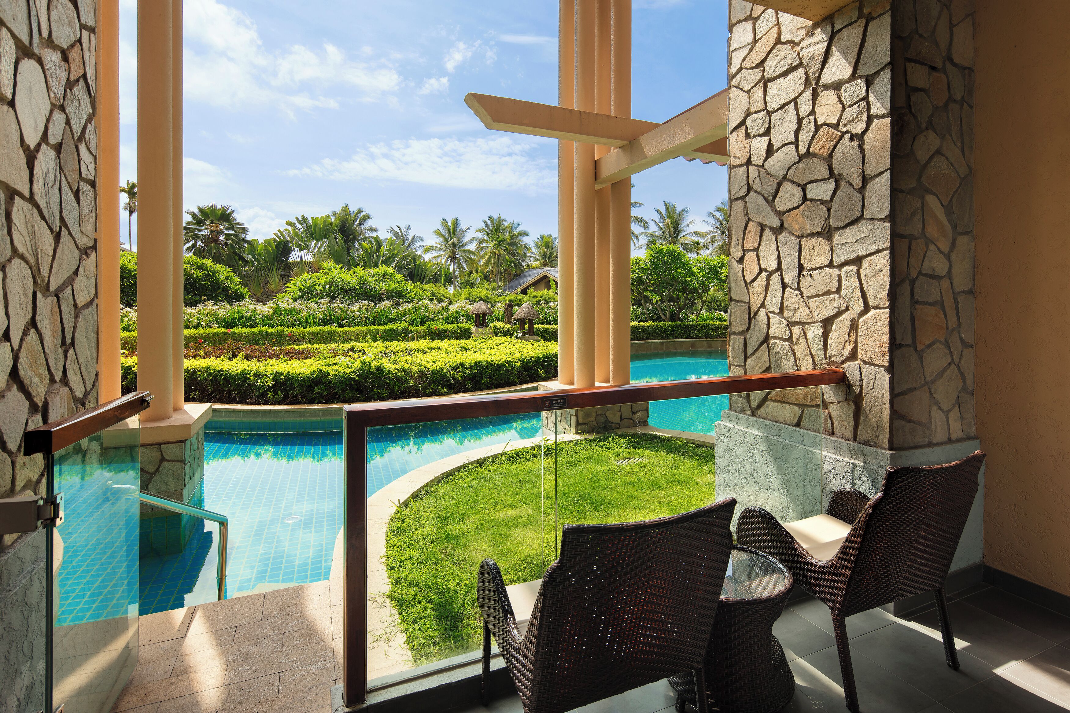 A poolside area with a glass railing, wicker chairs, and a stone pillar.