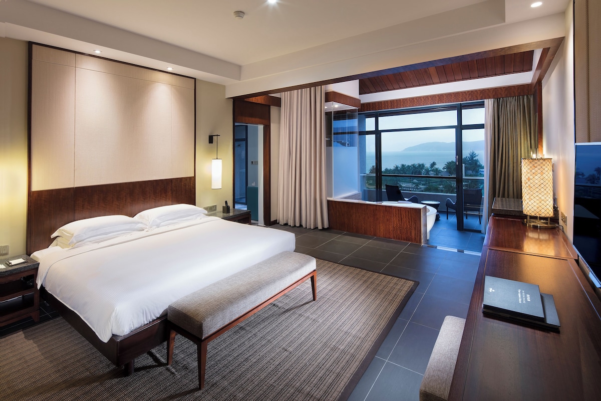 A modern hotel room with a large bed, a desk, a view of the ocean, and a balcony.