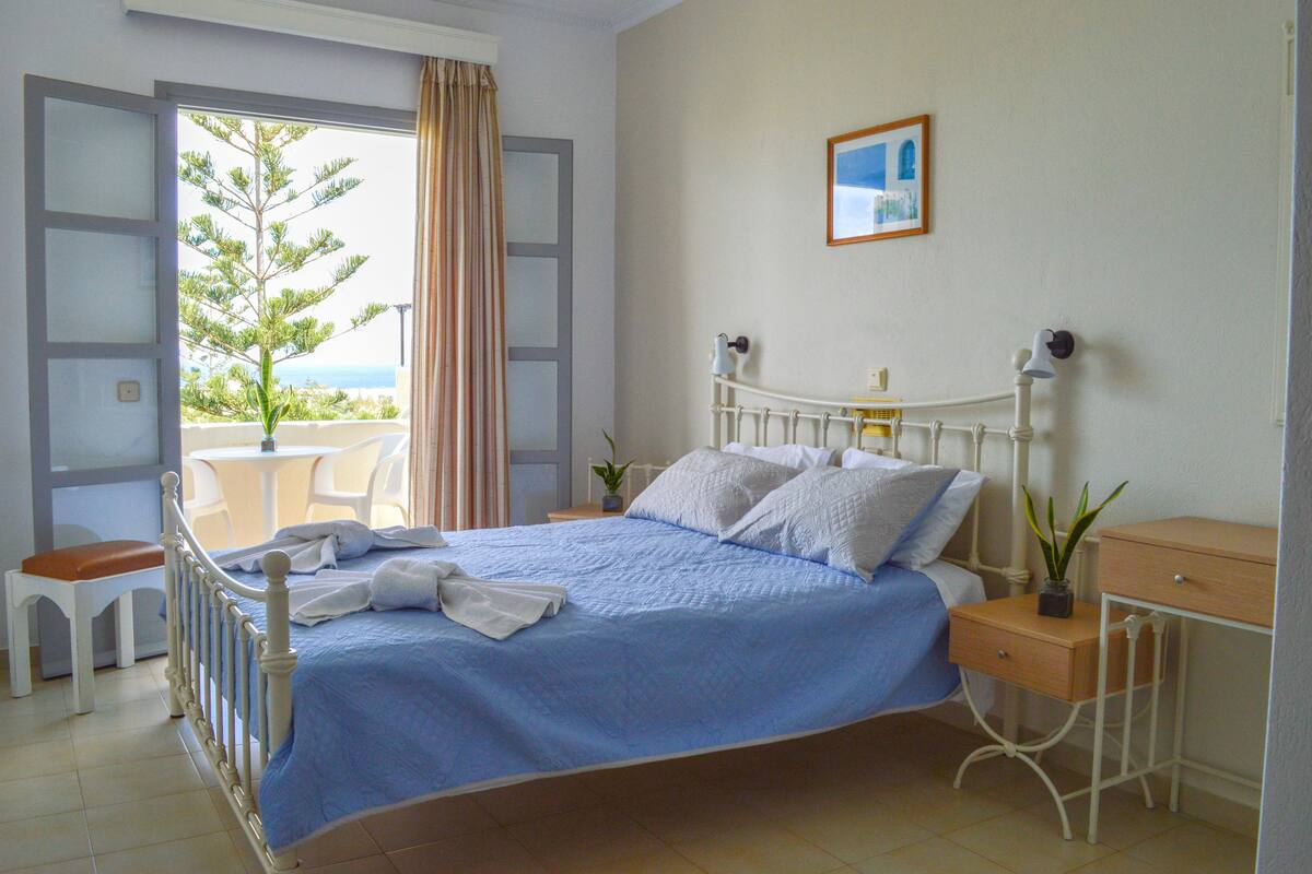 A bedroom with a bed, a bedside table, a nightstand, a window with curtains, and a view of the outdoors.