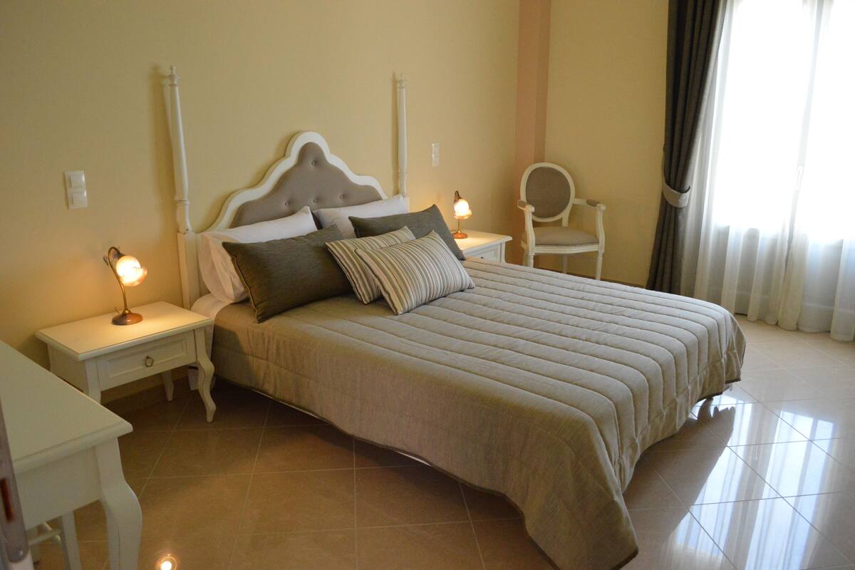 A neatly made bed with a striped pillow, a bedside lamp, and a chair in a well-lit room with a tiled floor.