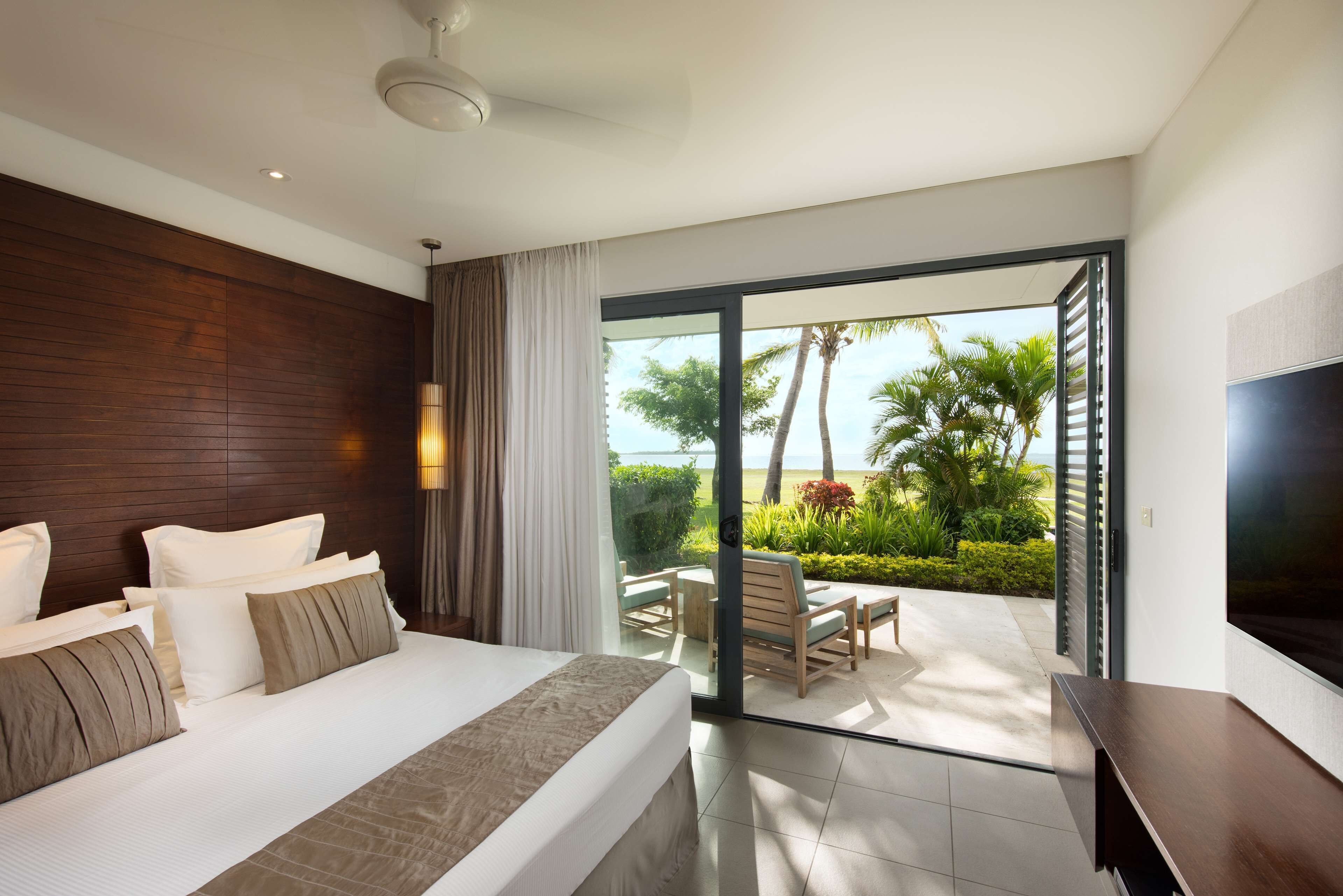 Una habitación de hotel moderna con una cama grande, televisión y balcón con vista a palmeras y al mar.