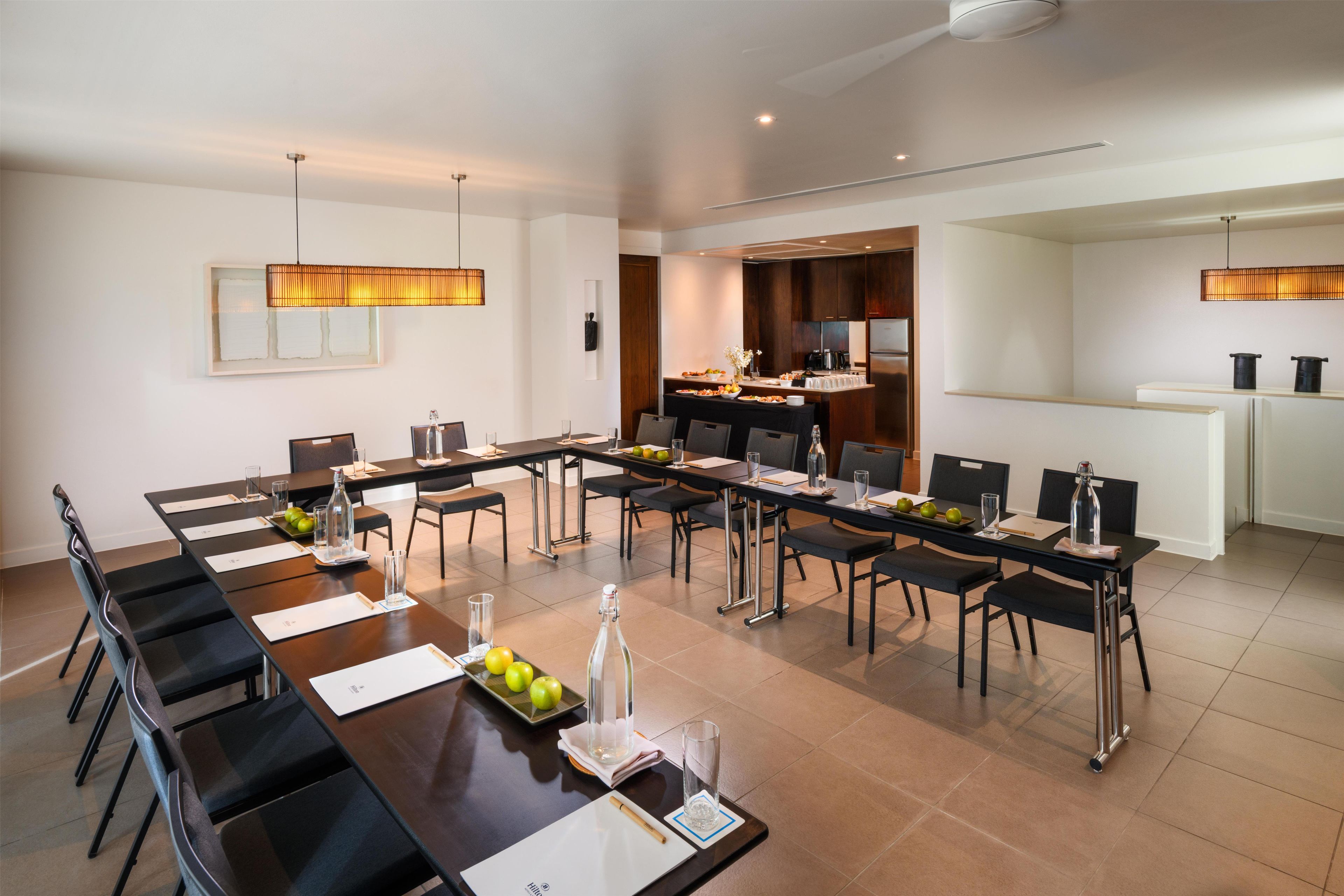 Un comedor moderno de hotel con una larga mesa común, asientos individuales y una cocina al fondo.