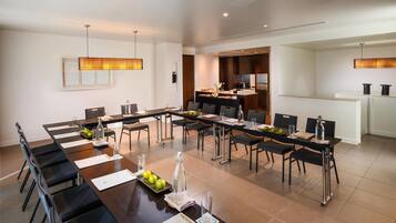 Un comedor moderno de hotel con una larga mesa común, asientos individuales y una cocina al fondo.