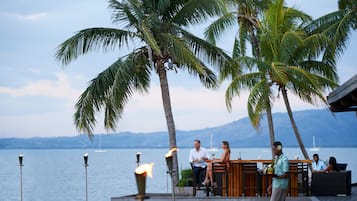 A waterfront bar with palm trees, a view of the sea, and a mountainous backdrop.