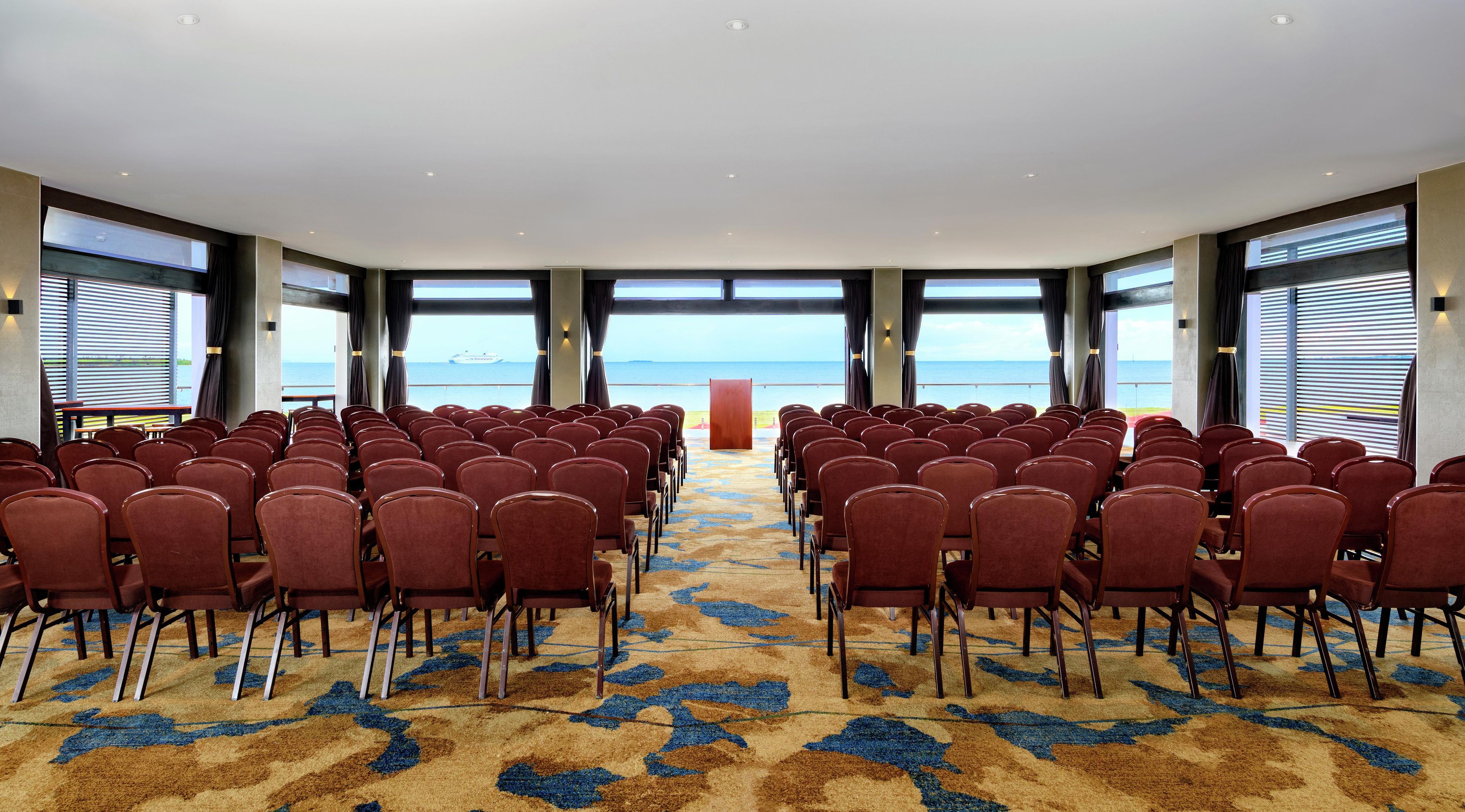 A conference room with rows of chairs facing a podium, large windows with a view of the sea, and a carpet with a blue and brown pattern.