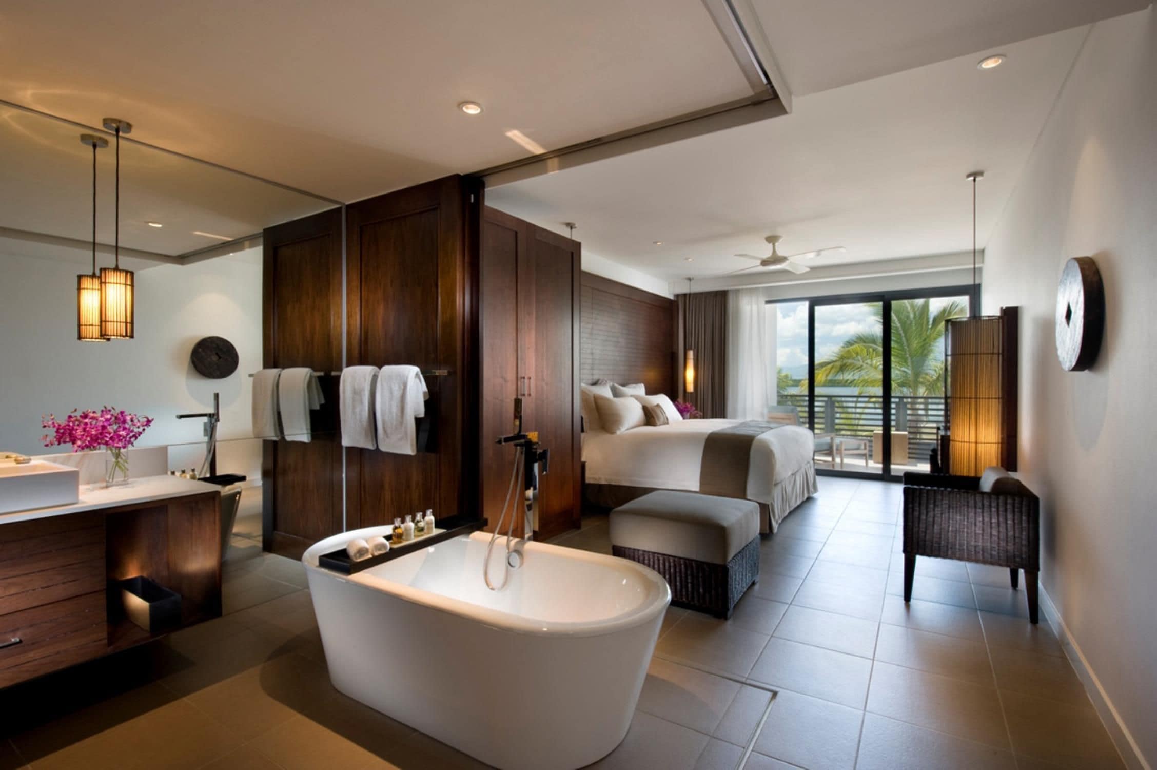 A modern hotel room with a large bathtub, a separate bedroom with a bed, a balcony with a view, and a bathroom with a vanity and towels.