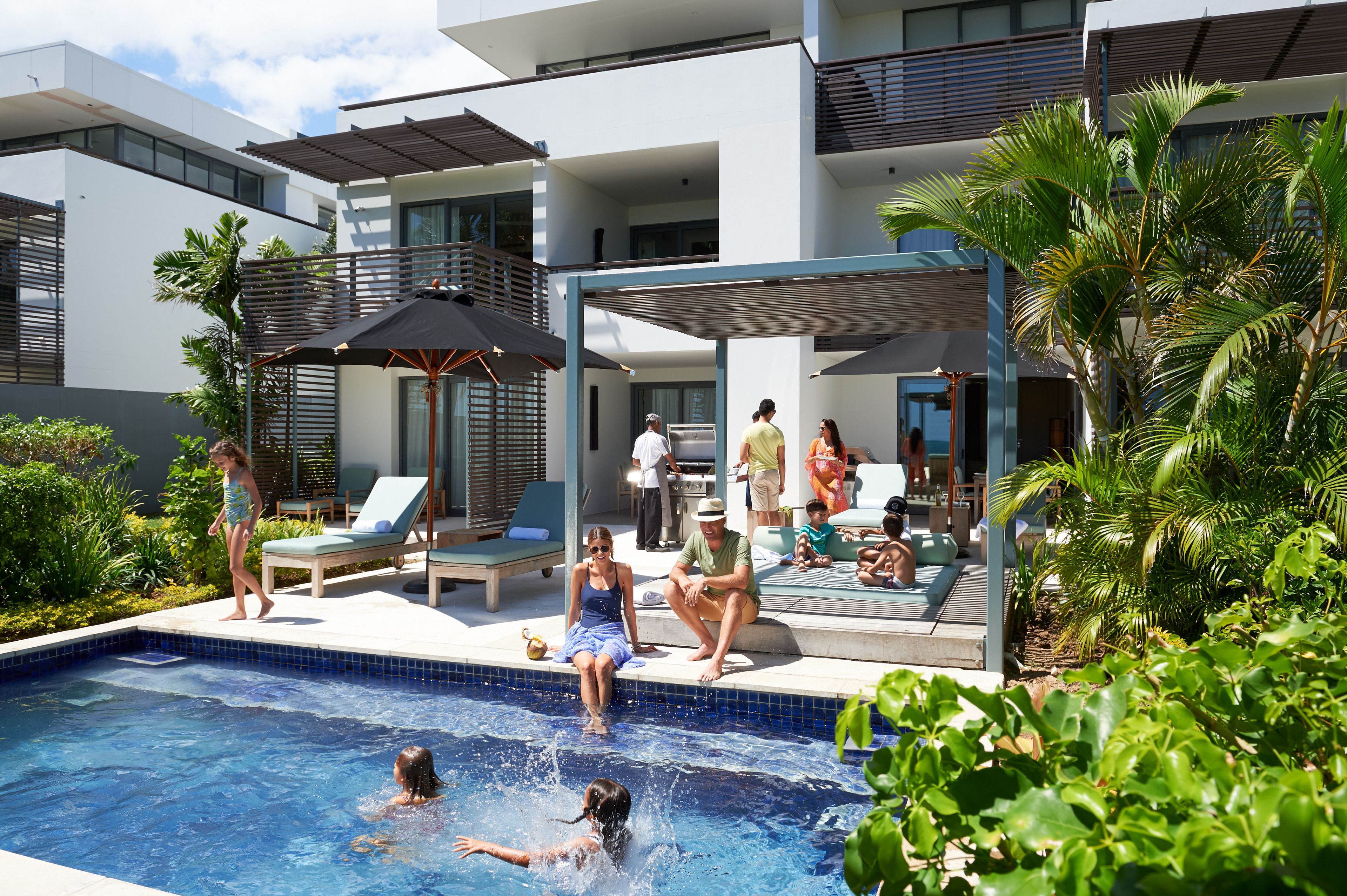 A modern outdoor pool area with lounge chairs and umbrellas, surrounded by greenery and a multi-level building.