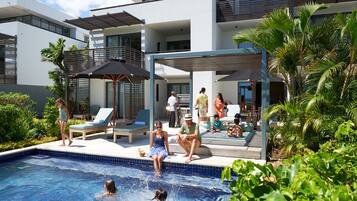 A modern outdoor pool area with lounge chairs and umbrellas, surrounded by greenery and a multi-level building.