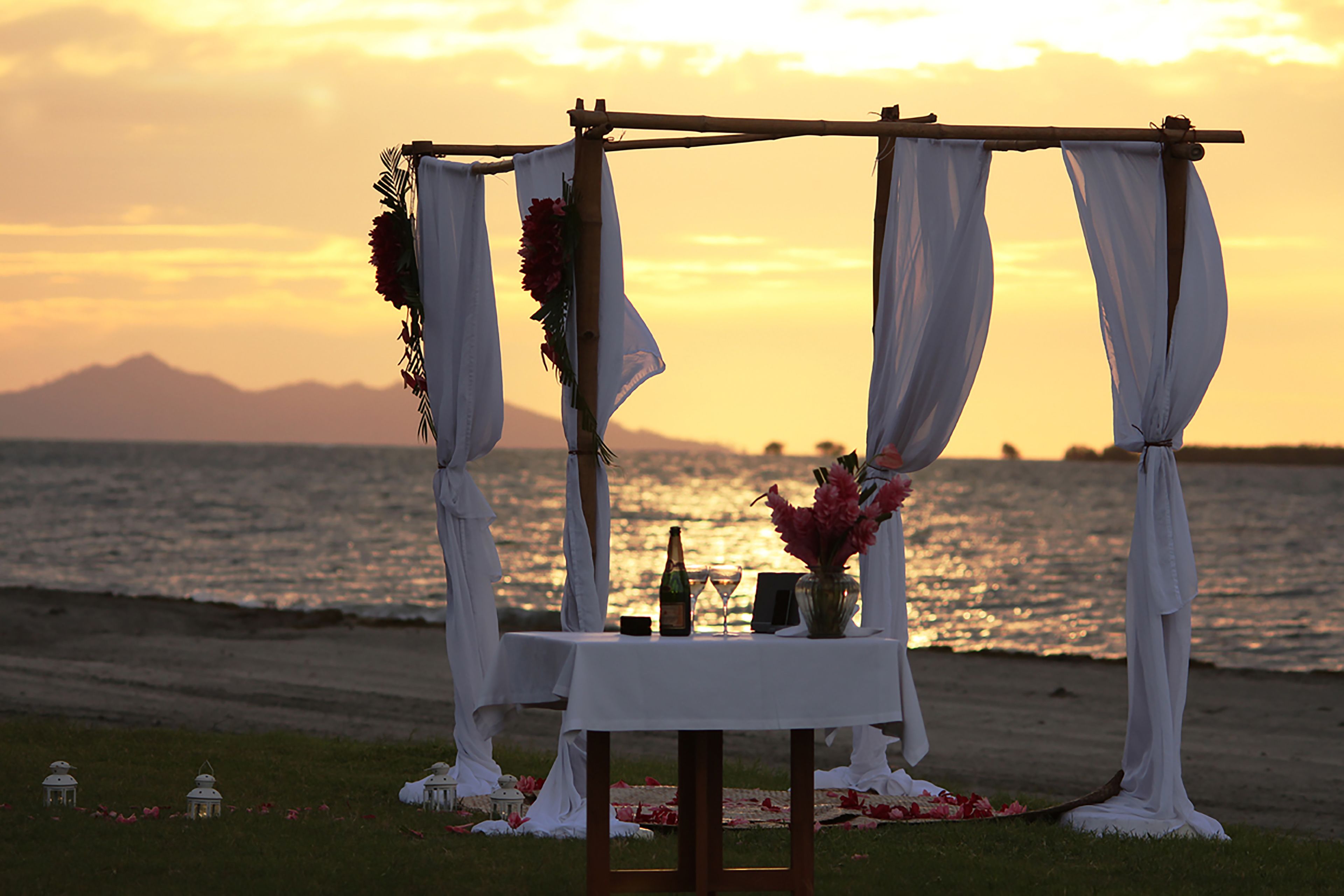 Un aménagement sur la plage avec une table, des draperies blanches et une bouteille, probablement pour un événement romantique.