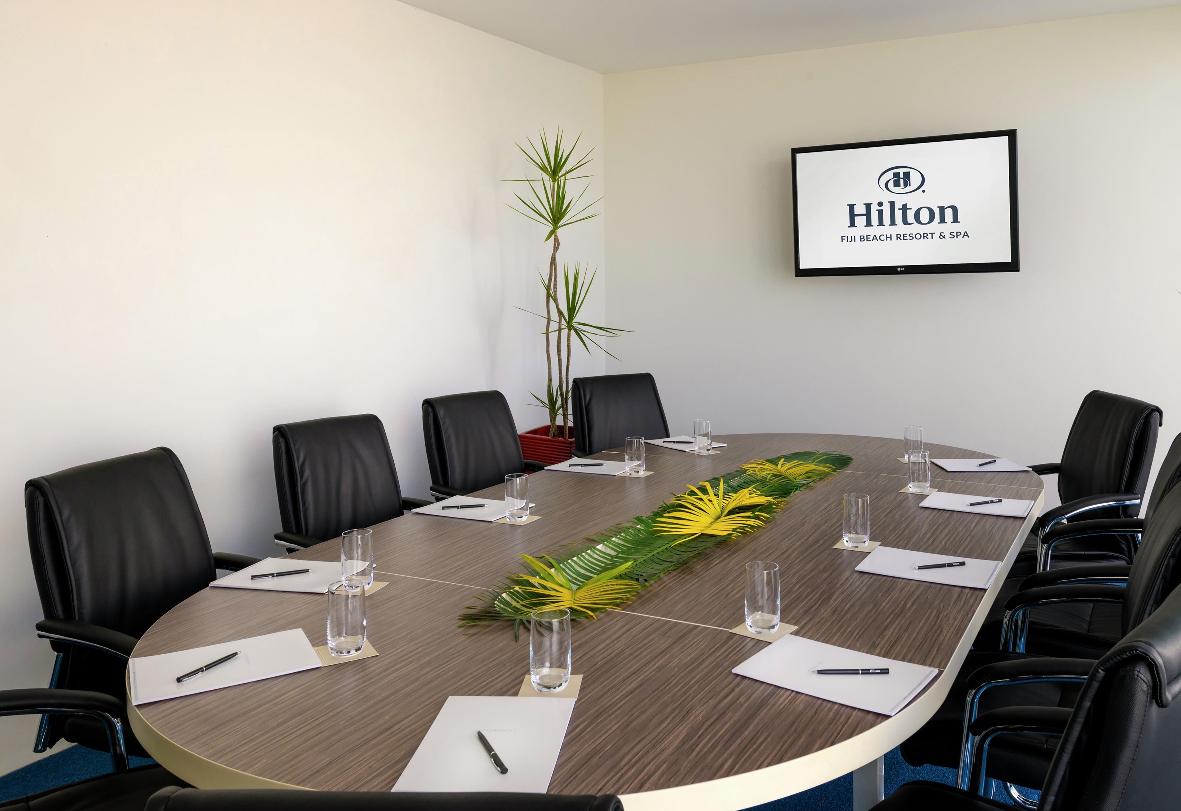 A conference room with a long table, black chairs, water glasses, pens, and a framed sign on the wall.
