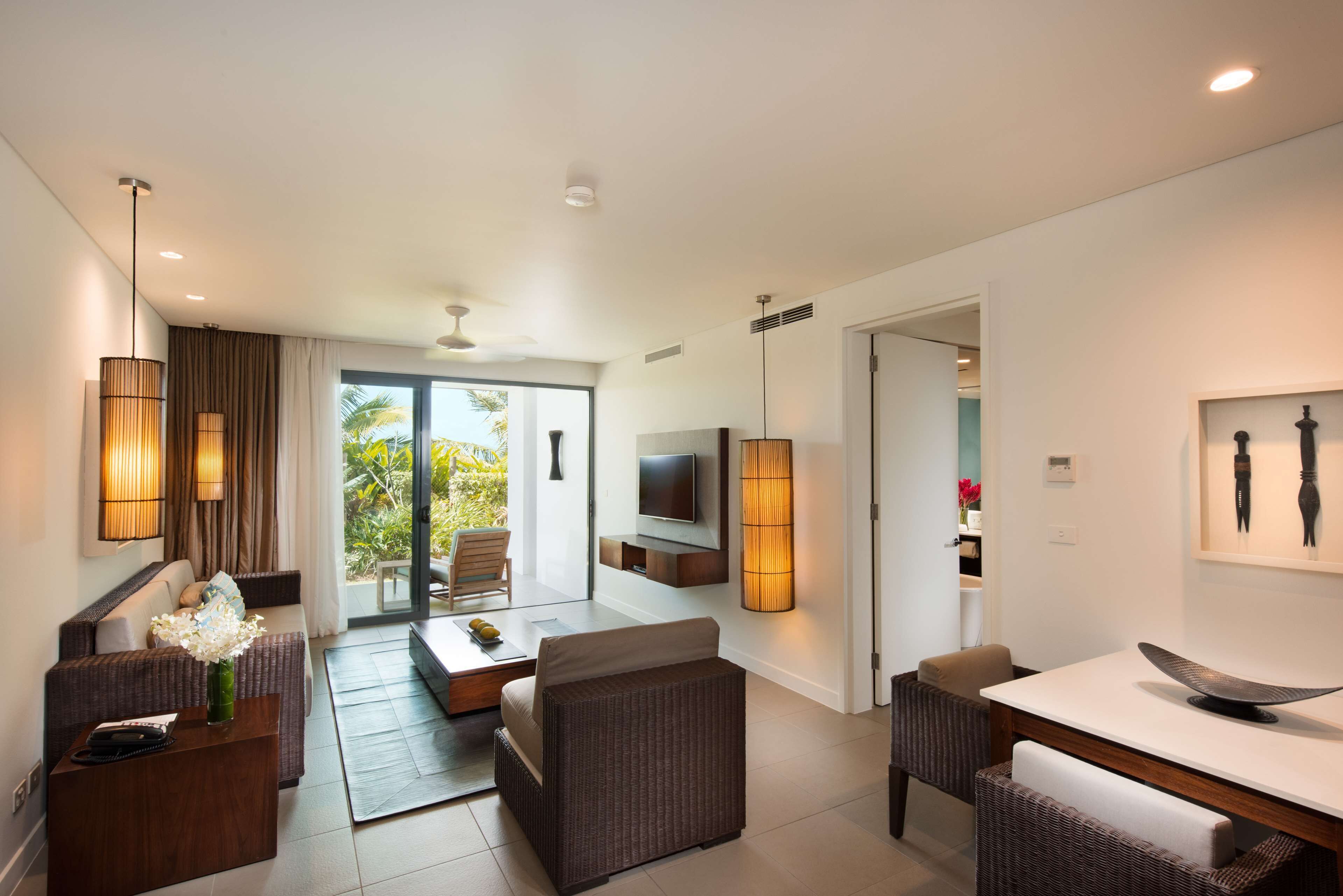 Habitación de hotel moderna con sofá, mesa de comedor y televisión. Tiene un balcón con vista a palmeras.