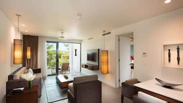 Habitación de hotel moderna con sofá, mesa de comedor y televisión. Tiene un balcón con vista a palmeras.