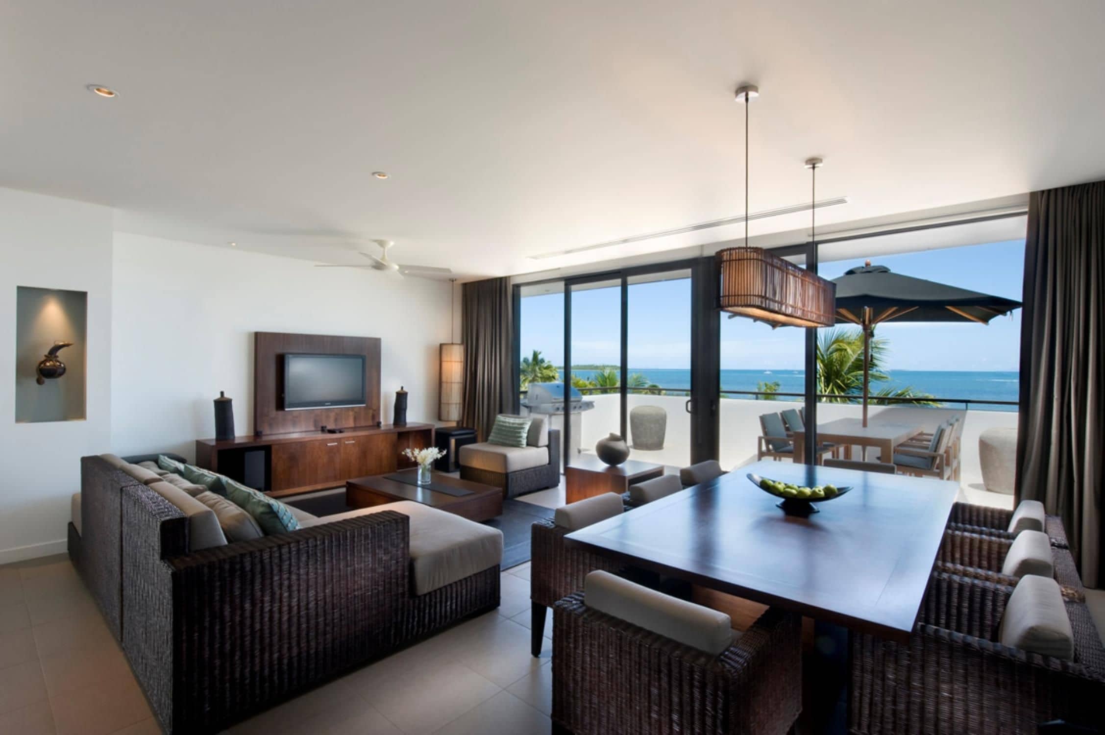 A modern living room with a flat-screen TV, comfortable seating, a dining area, and a large window offering a view of the ocean.