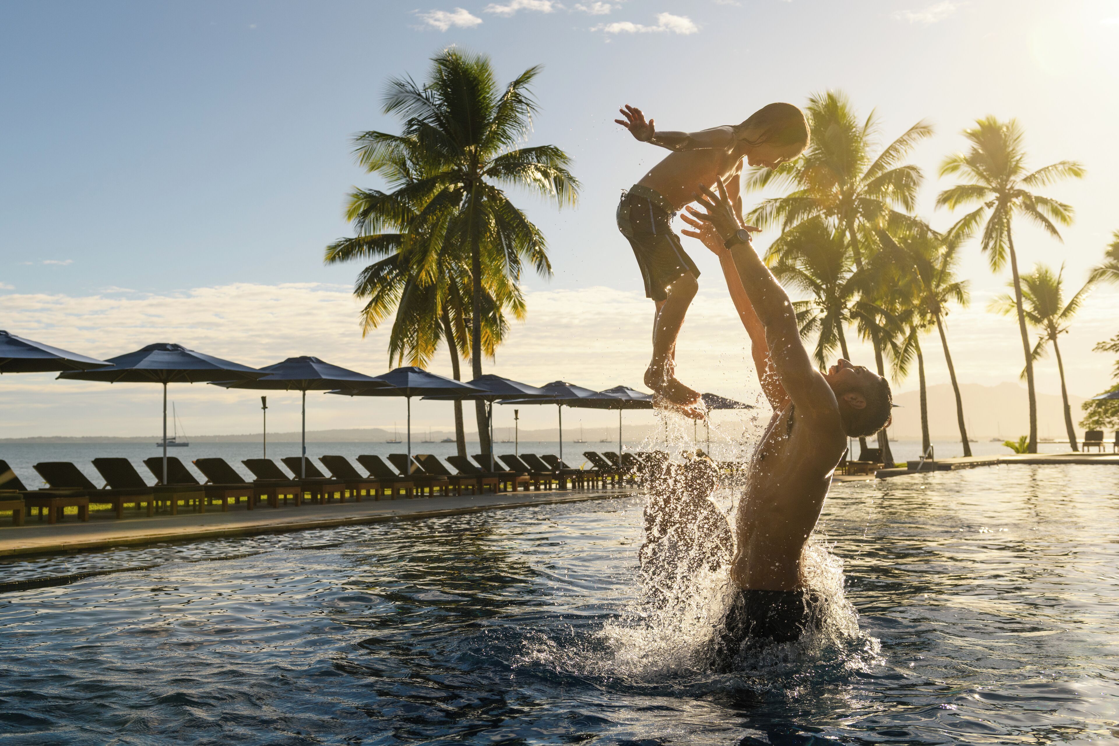 Una persona salta a una piscina, con palmeras y sombrillas de fondo.