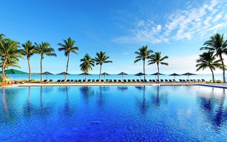 A pool with lounge chairs and umbrellas, palm trees, and a clear sky.