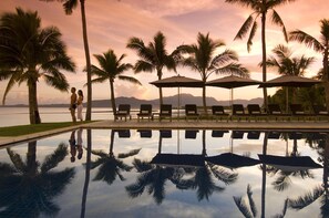 A poolside area with lounge chairs and umbrellas, palm trees, and a view of the ocean and mountains.