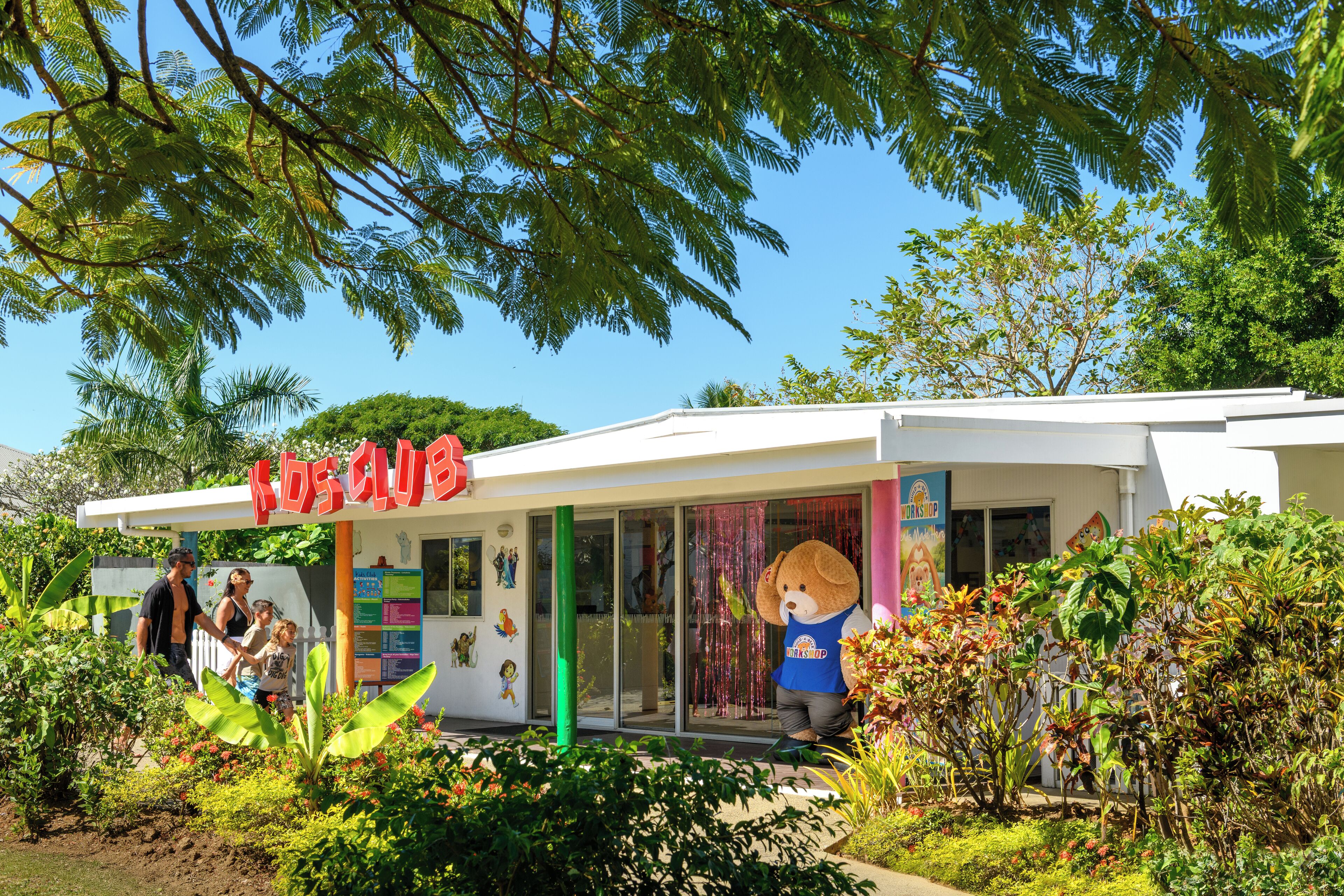 A children's play area with a large teddy bear statue, a white building, and lush greenery.