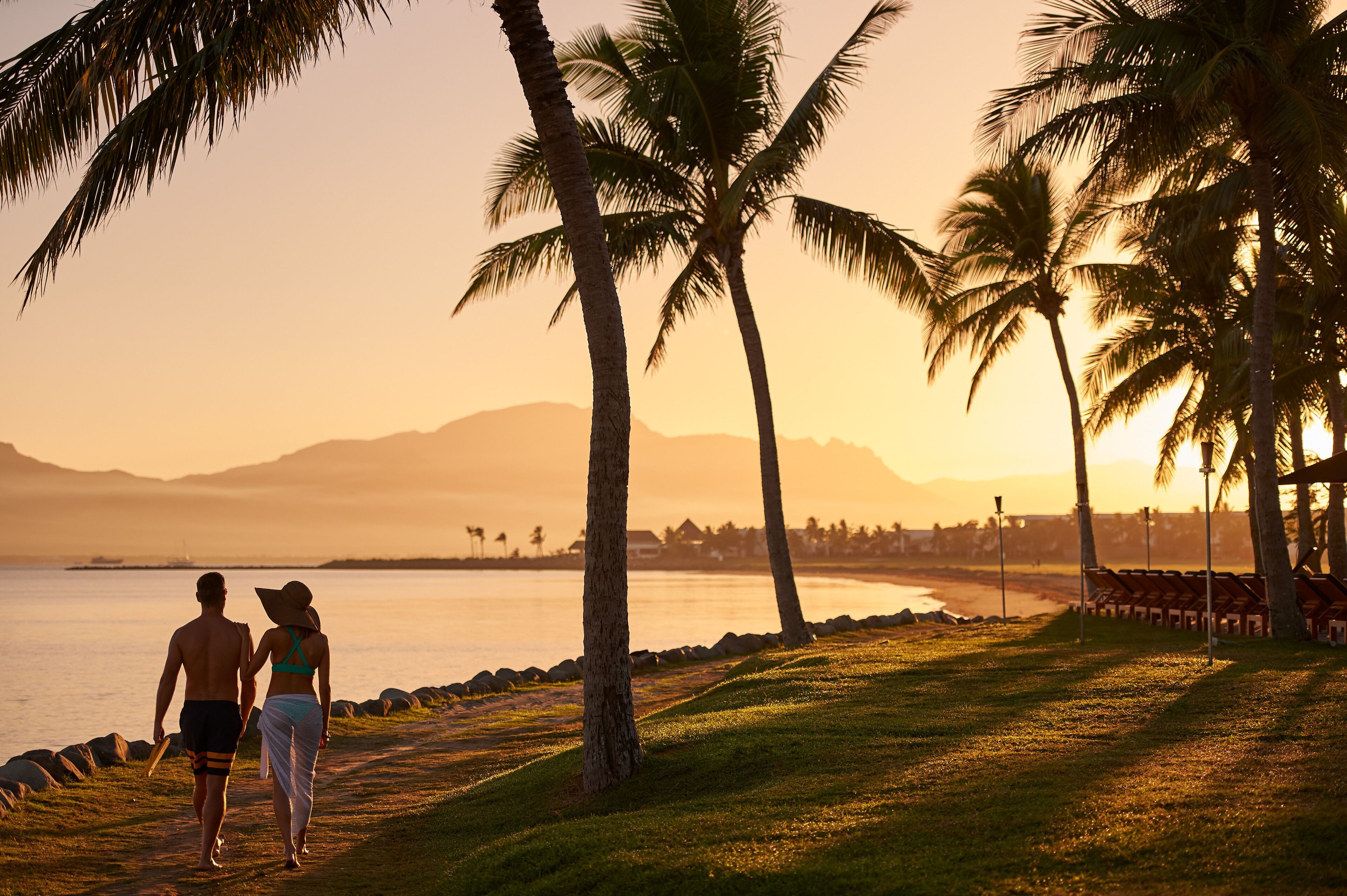 A couple walking along a beach path with palm trees and a sunset in the background.