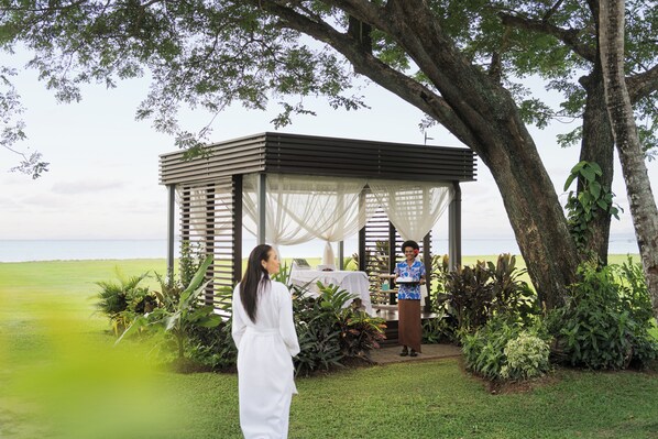 A gazebo with a table and chairs, surrounded by greenery and a view of the sea.