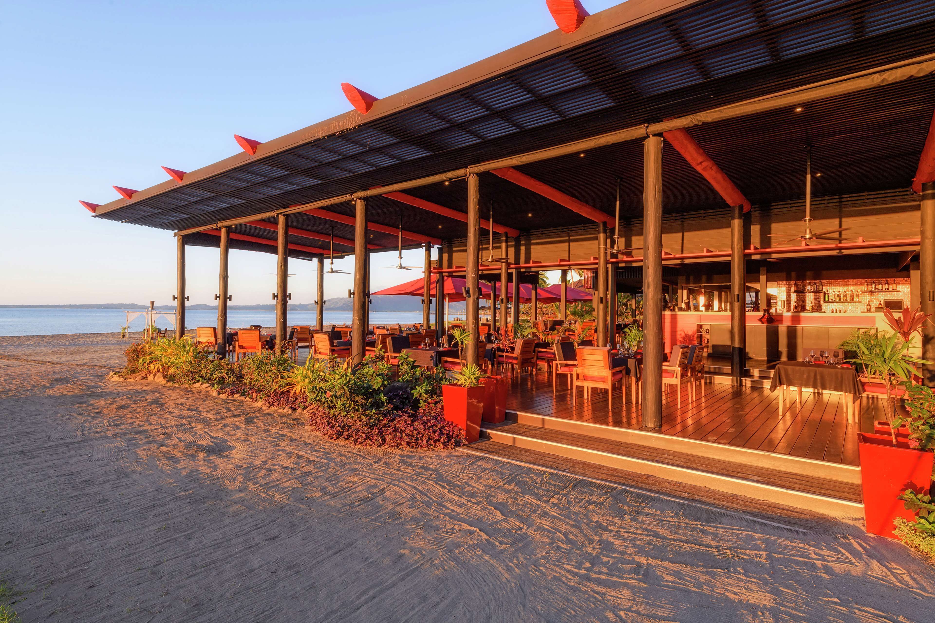 A beachfront restaurant with outdoor seating, a bar, and a view of the ocean.