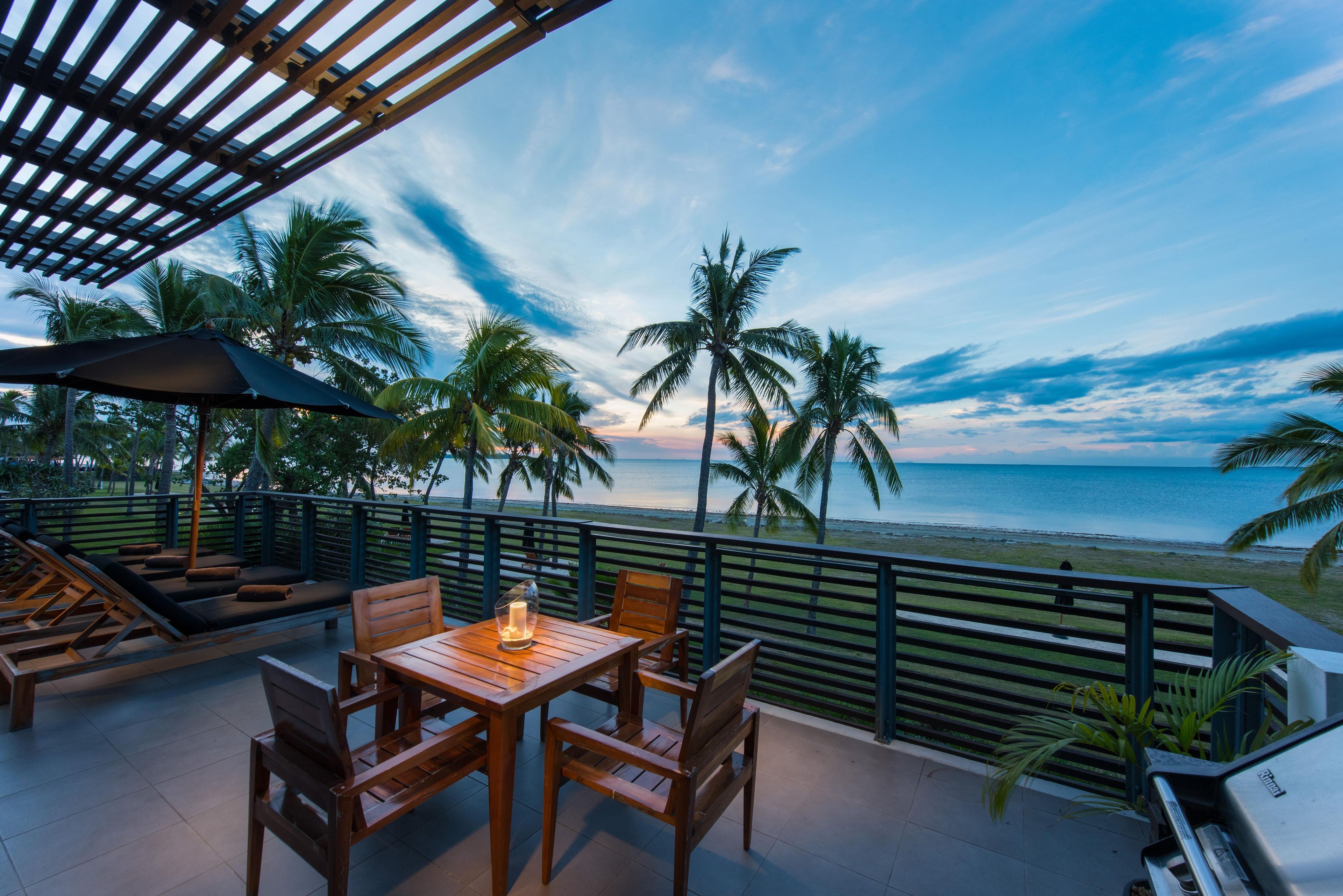 An outdoor seating area with a table and chairs, overlooking a beach with palm trees and a view of the ocean.