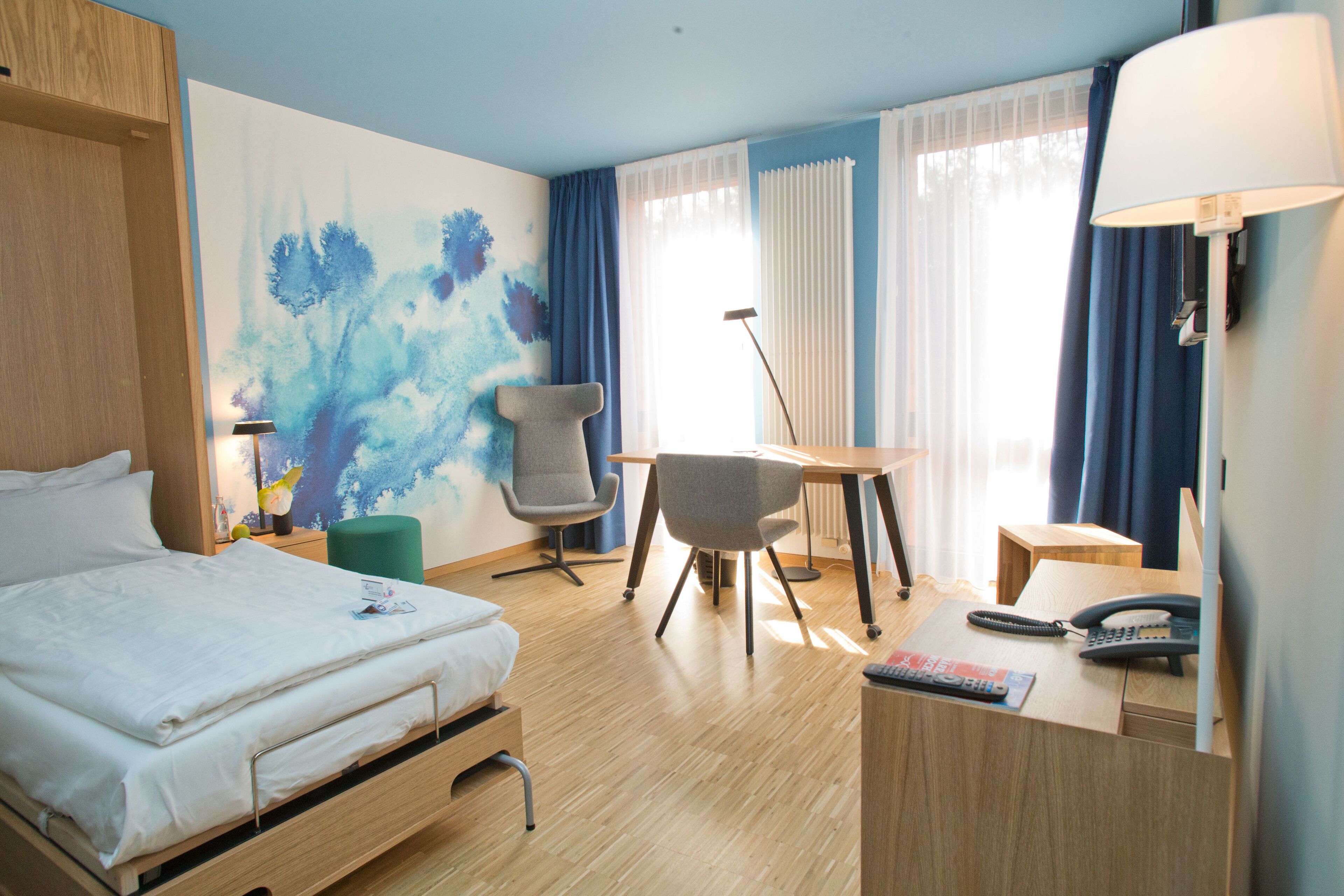 Une chambre d’hôtel avec un lit, un bureau et une chaise, une lampe, un téléphone et un tableau au mur.
