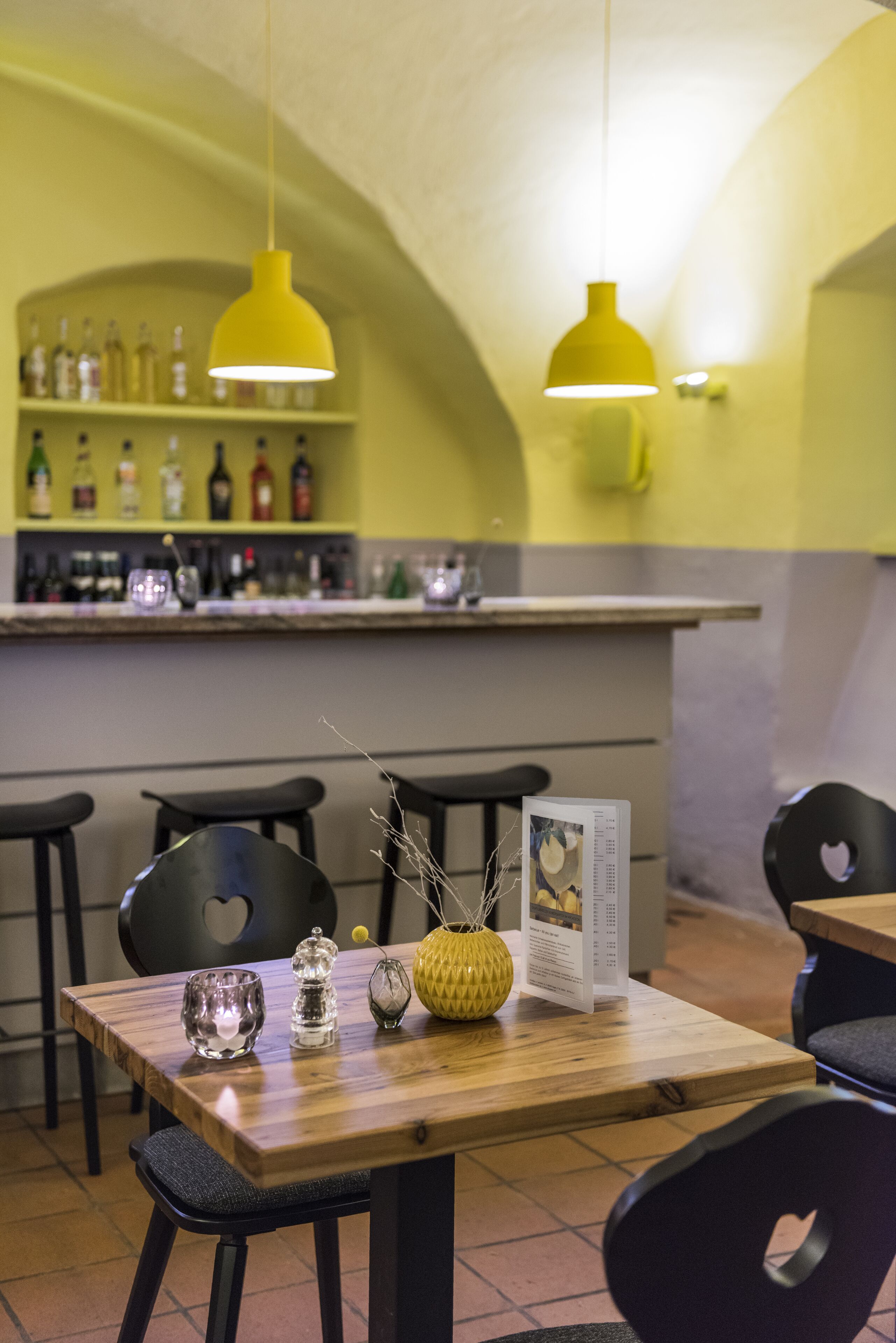 A restaurant interior with a wooden table, black chairs, yellow pendant lights, and a bar area with bottles and glasses.