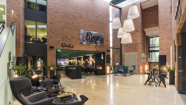 A spacious lobby with a fireplace, seating areas, and a visible restaurant named 'Emils'. - Arcadeon (Hagen)