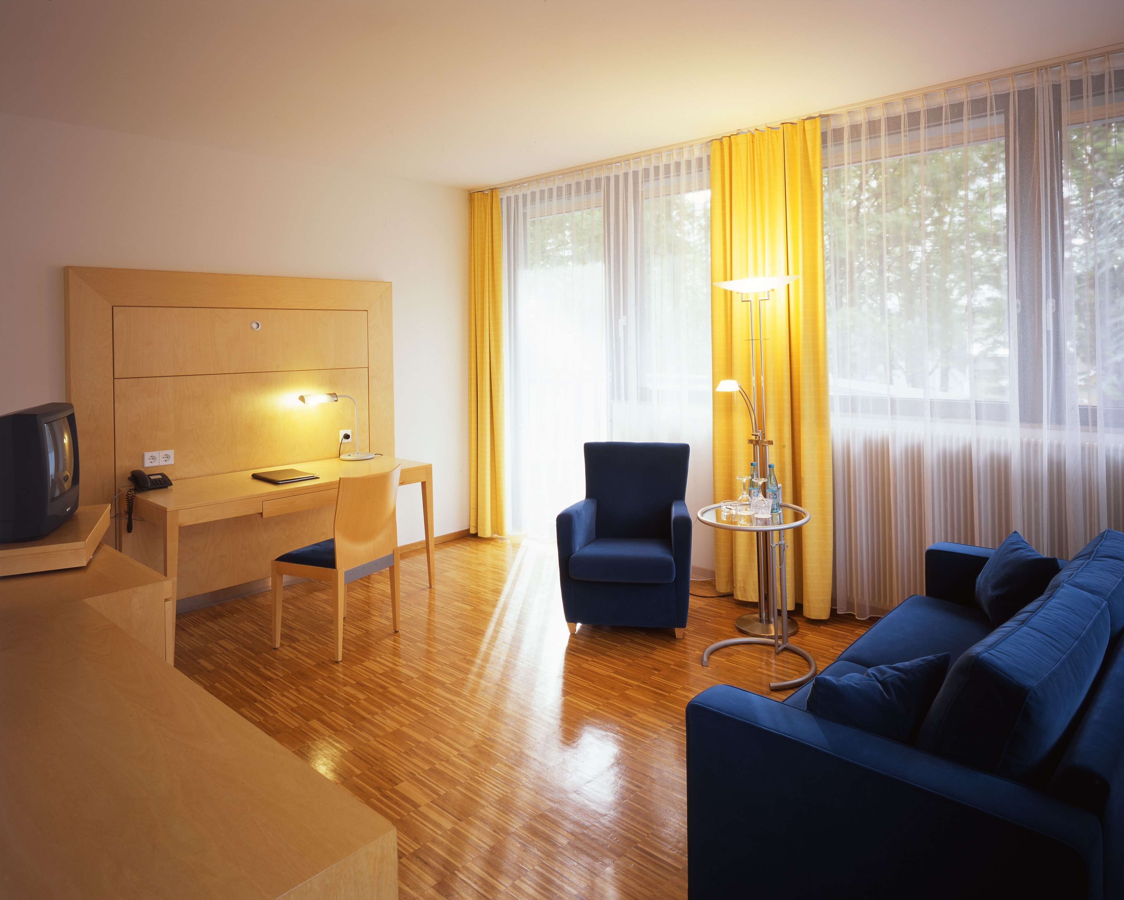 Ein Hotelzimmer mit einem Holztisch, einem blauen Sessel, einem blauen Sofa, einer Stehlampe und einem Fernseher.