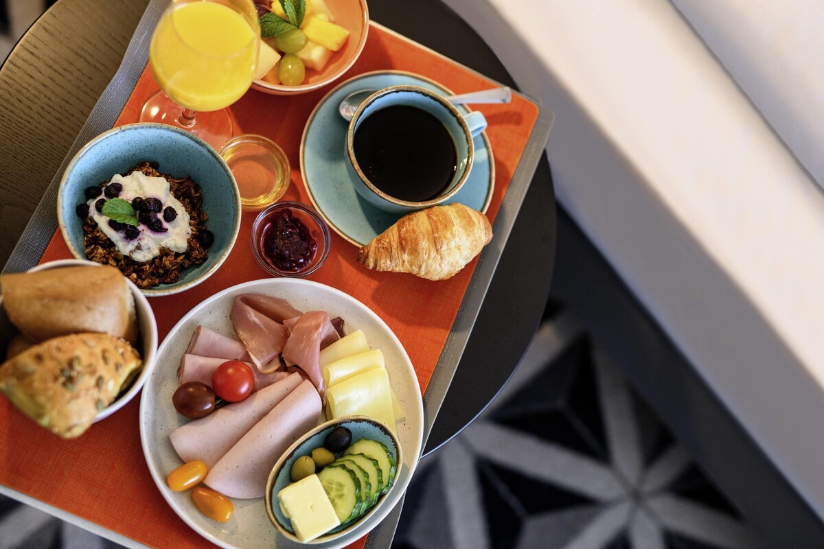 Bandeja de desayuno con diversas opciones de comida, incluyendo cruasanes, queso, jamón, tomates, aceitunas y una cesta de frutas con uvas y piña.