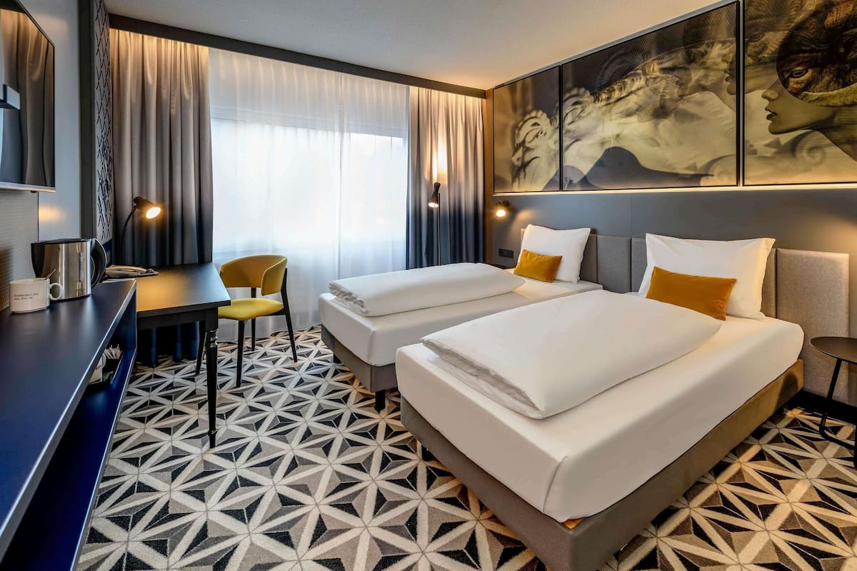 A hotel room with two beds, a desk, a chair, a TV, and a geometric patterned floor.