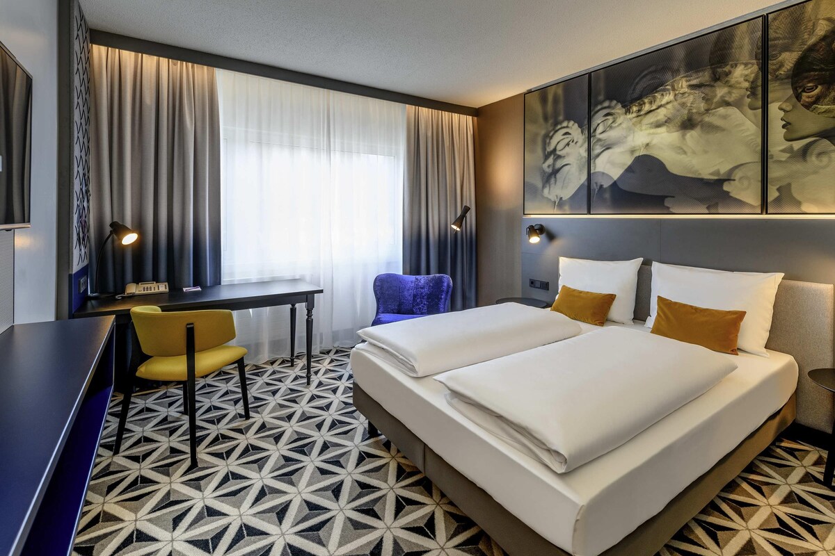 Une chambre d’hôtel comprenant un lit, un bureau, une chaise, une grande fenêtre avec des rideaux et un sol orné d’un motif géométrique.