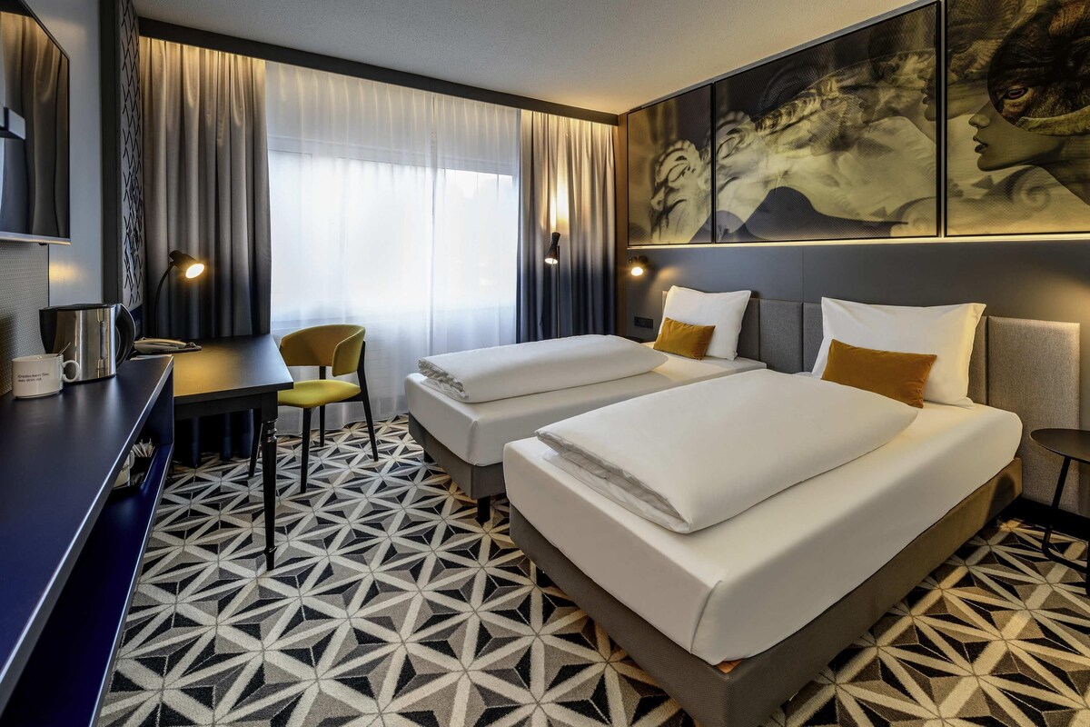 Habitación de hotel con dos camas, un escritorio, una silla y un cuadro grande en la pared.