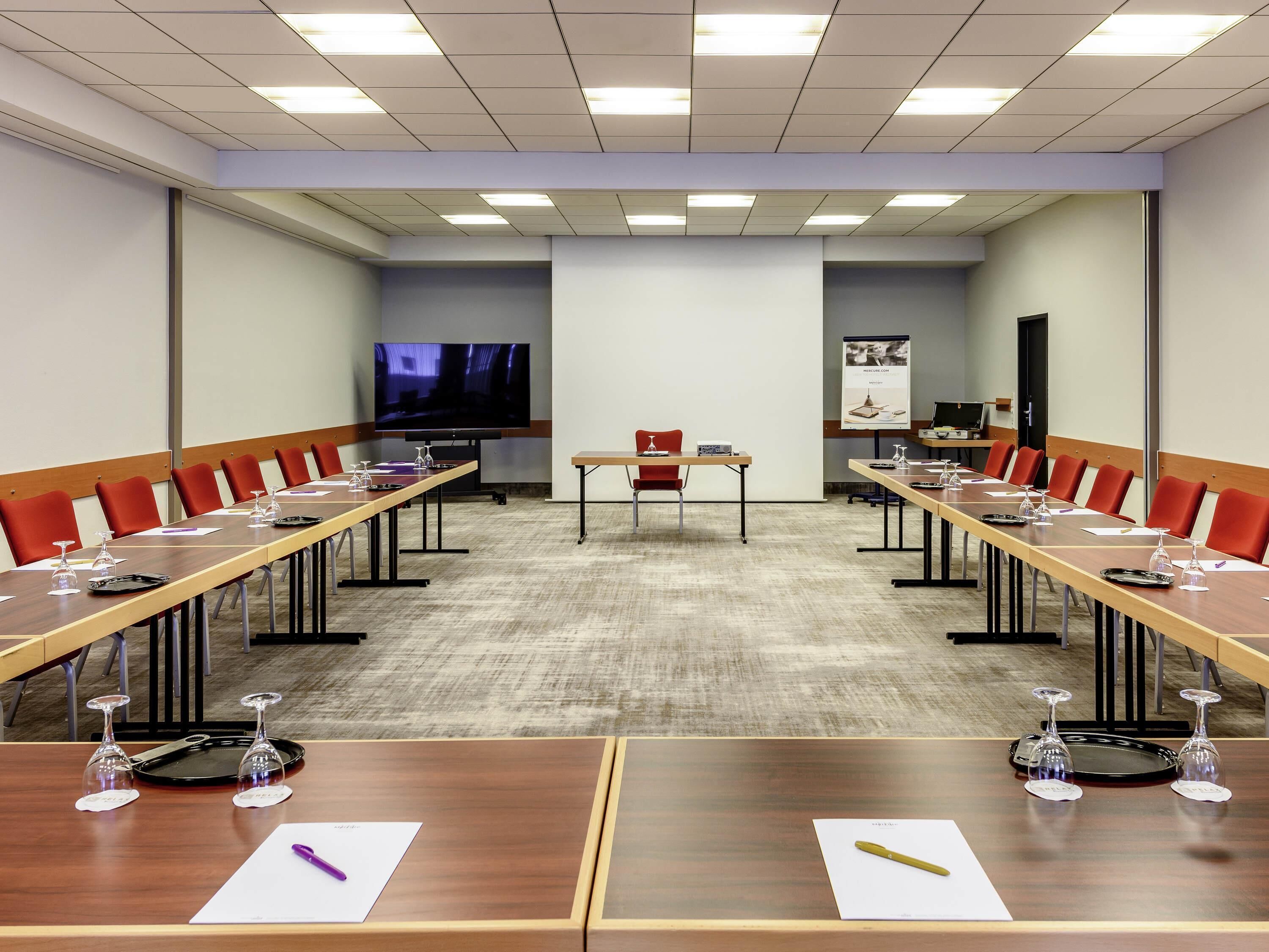 A conference room with long tables, red chairs, a large screen, and a poster on the wall.