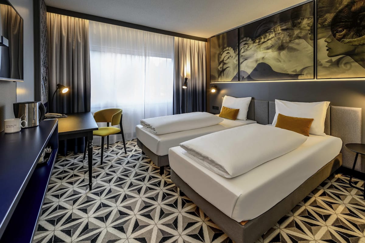 A hotel room with two beds, a desk, a chair, and a large painting on the wall.