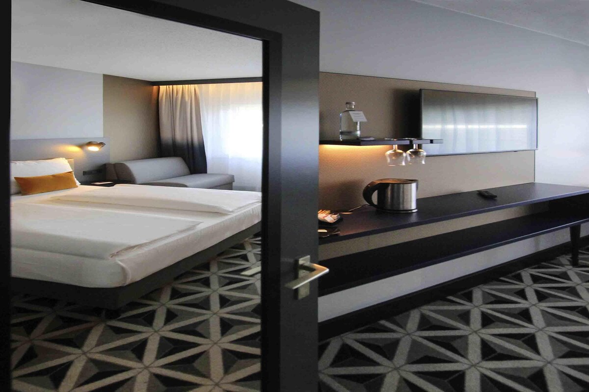 Habitación de hotel con cama, escritorio, espejo, hervidor de agua y piso estampado.