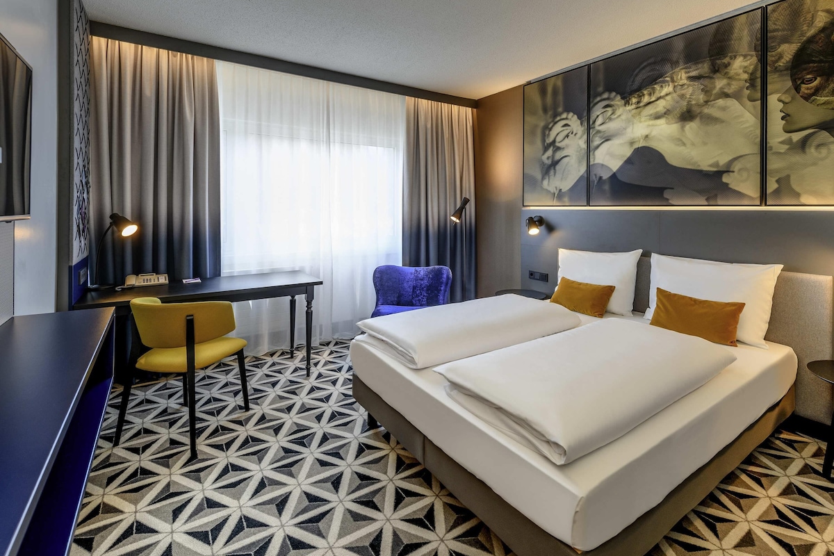 Habitación de hotel con cama, escritorio, silla, ventana grande con cortinas y piso con diseño geométrico.