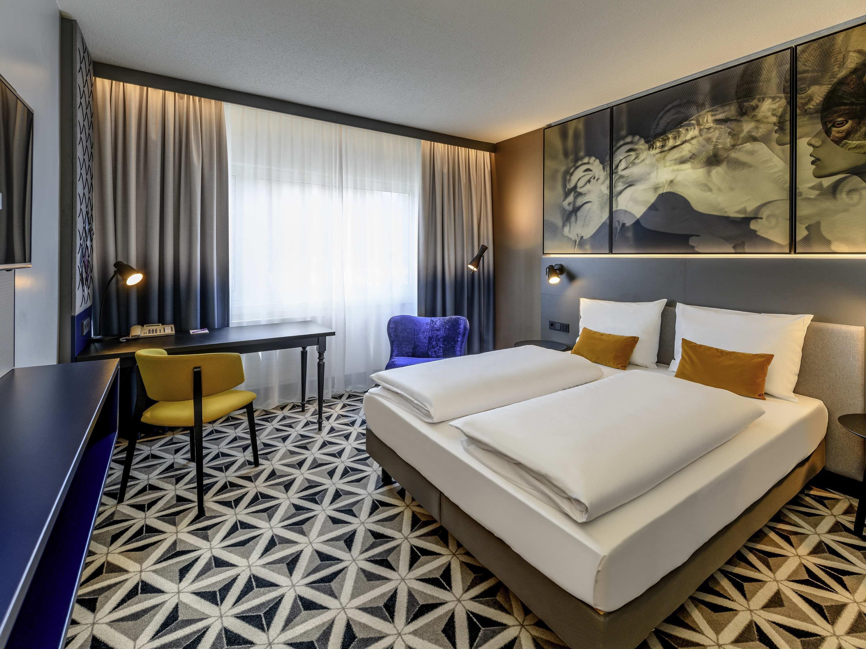 A hotel room with a bed, a desk, a chair, a large window with curtains, and a geometric patterned floor.